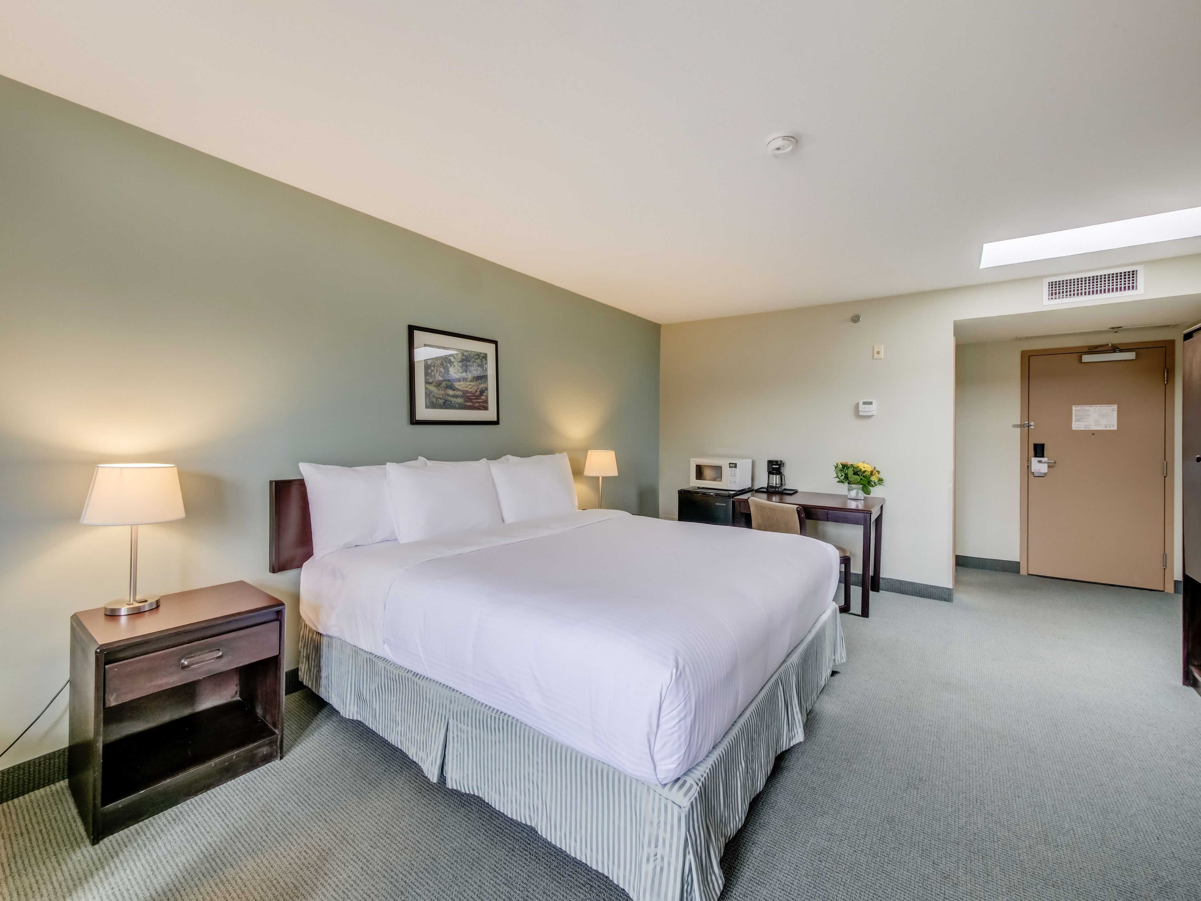 Habitación de hotel con una cama grande, mesitas de noche con lámparas, un escritorio con computadora, un microondas y una puerta con un letrero.