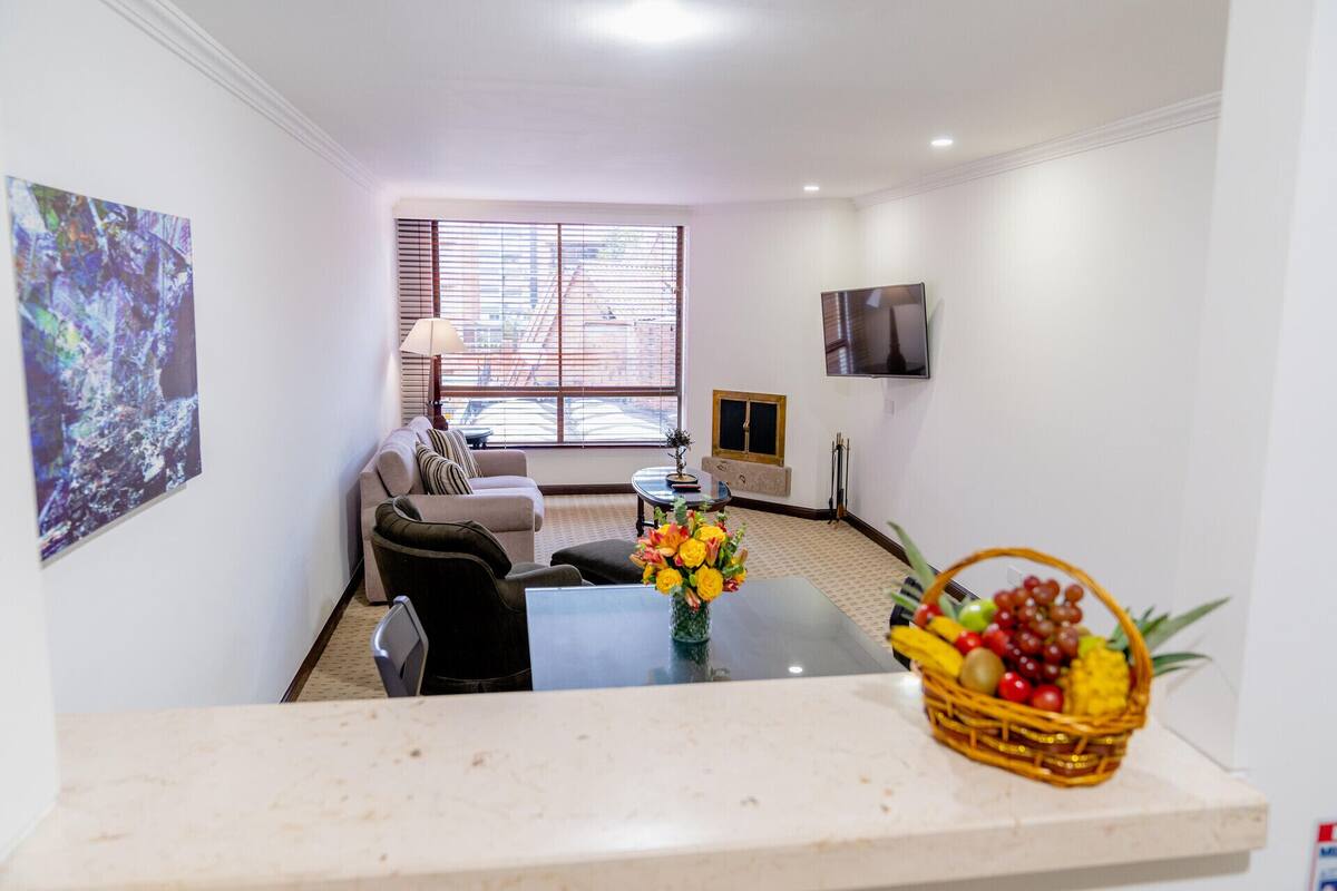 A living room with a sofa, a coffee table with a fruit basket, a lamp, and a TV on the wall.