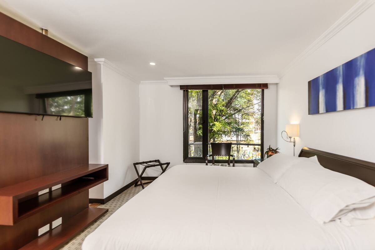 A hotel room with a large bed, a TV mounted on the wall, a balcony with a chair, and a view of greenery outside.