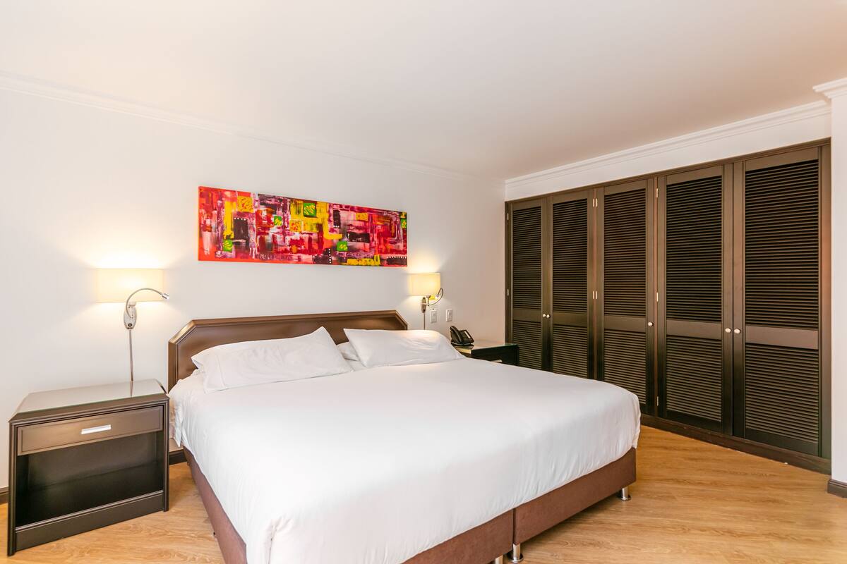Cama bem arrumada em quarto de hotel com uma grande obra de arte na parede, abajur de cabeceira e guarda-roupa com portas venezianas.