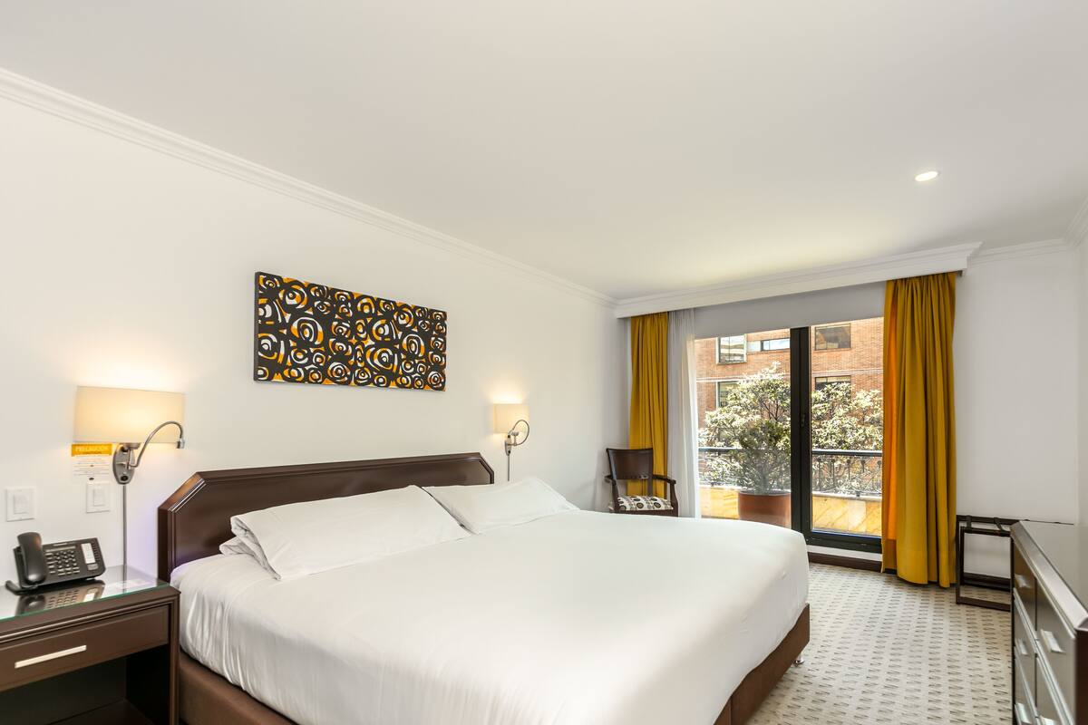 Quarto de hotel com uma cama grande, uma mesa com telefone, uma cadeira, uma janela com cortinas amarelas e vista para o exterior.