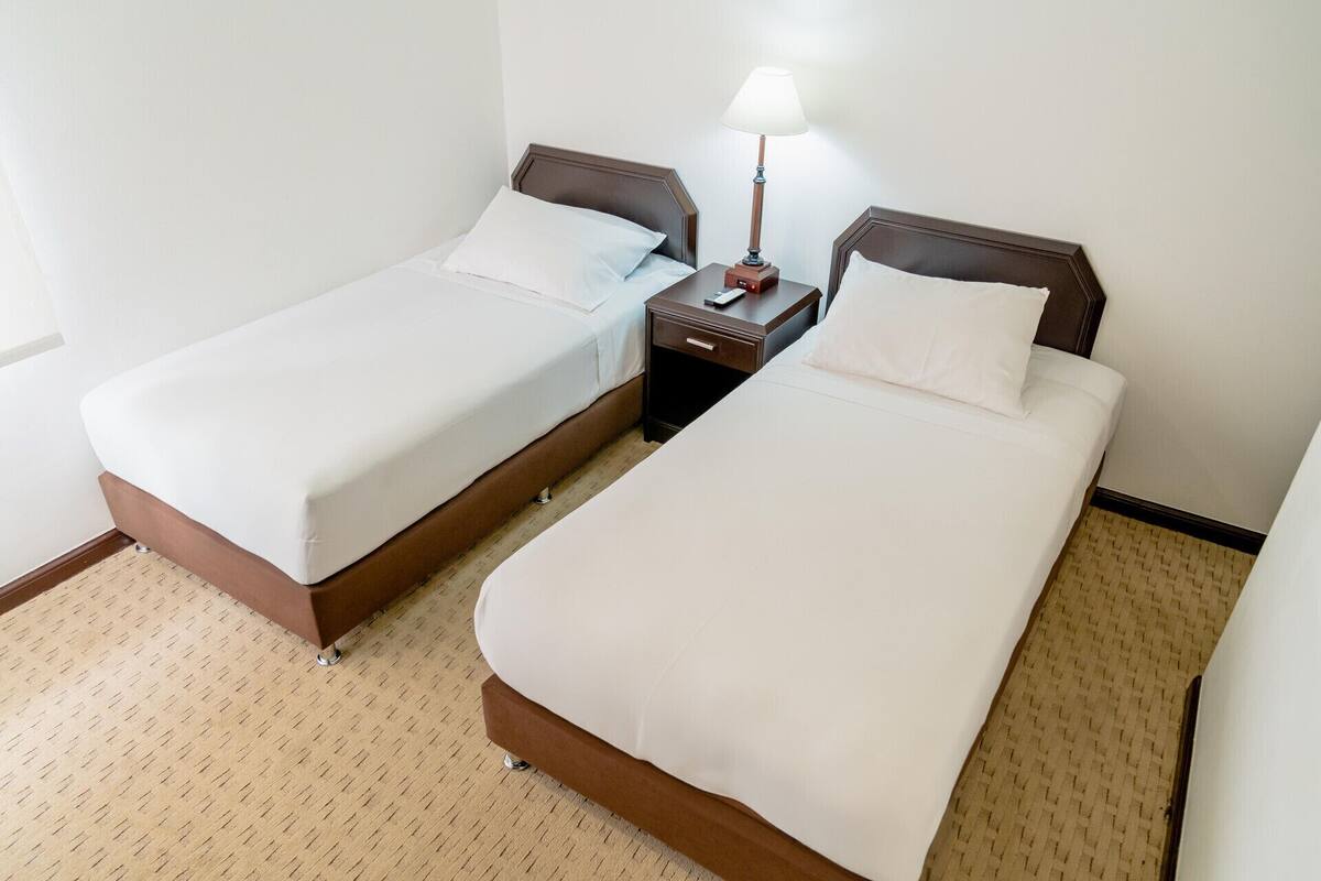 A hotel room with two single beds, a nightstand with a lamp, and a carpeted floor.