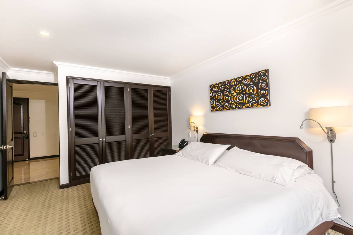A hotel room with a large bed, a wardrobe, a bedside table with a lamp, and a wall-mounted artwork.