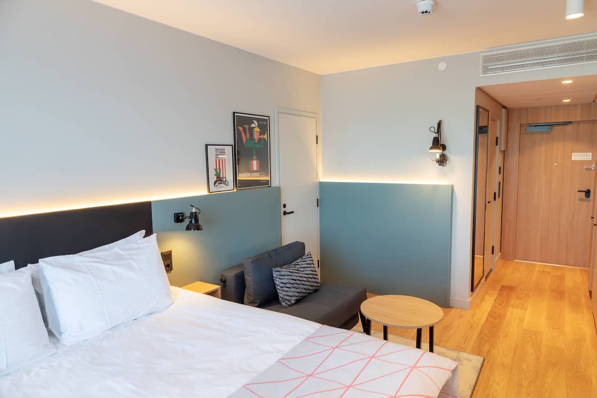 A hotel room with a bed, a sofa, a small table, and a wall-mounted lamp.