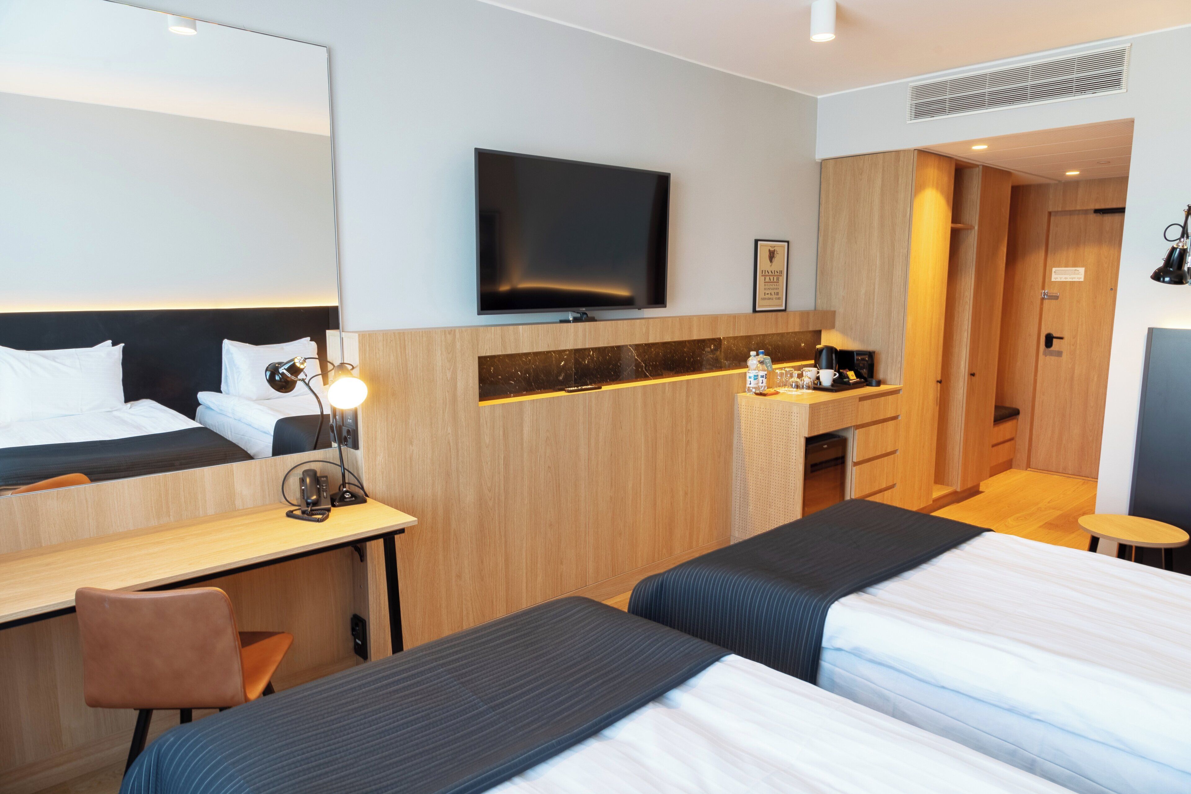 Habitación de hotel con dos camas, un escritorio, un espejo, una televisión y una pequeña kitchenette.