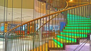 A spiral staircase with a wooden handrail and metal railings, leading to a brightly lit area with green illuminated steps.