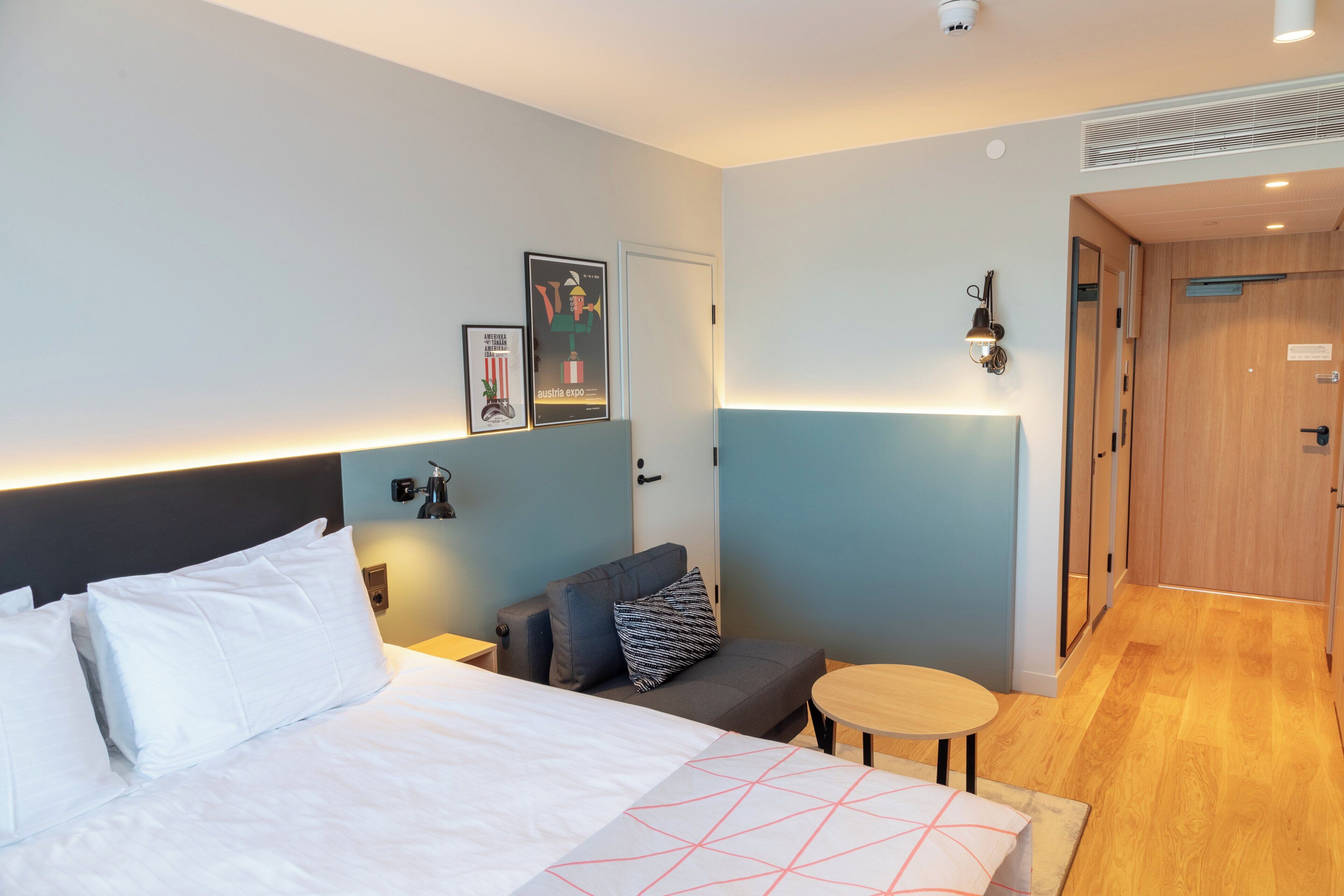 Una camera d'albergo con un letto, un tavolino, una poltrona e una porta che dà su un'altra stanza.