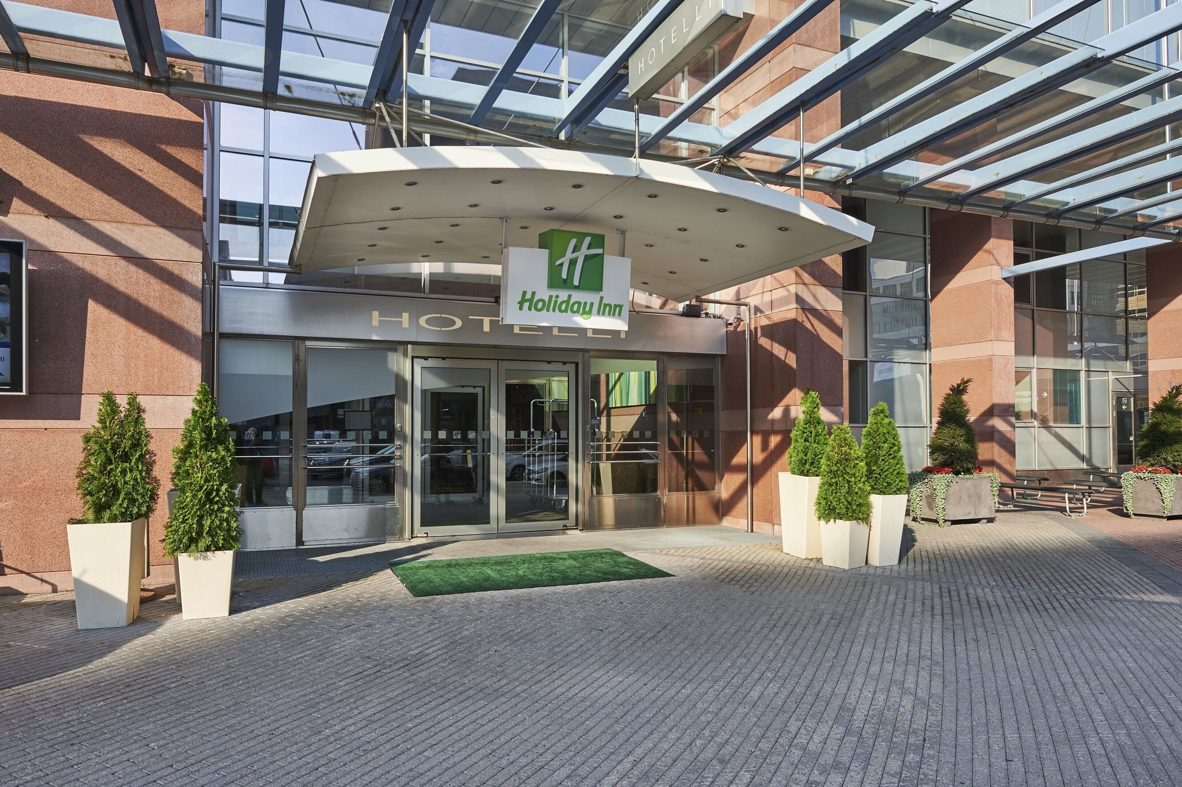 Entrada de um hotel Holiday Inn, com marquise de vidro, placa de identificação e plantas em vasos.