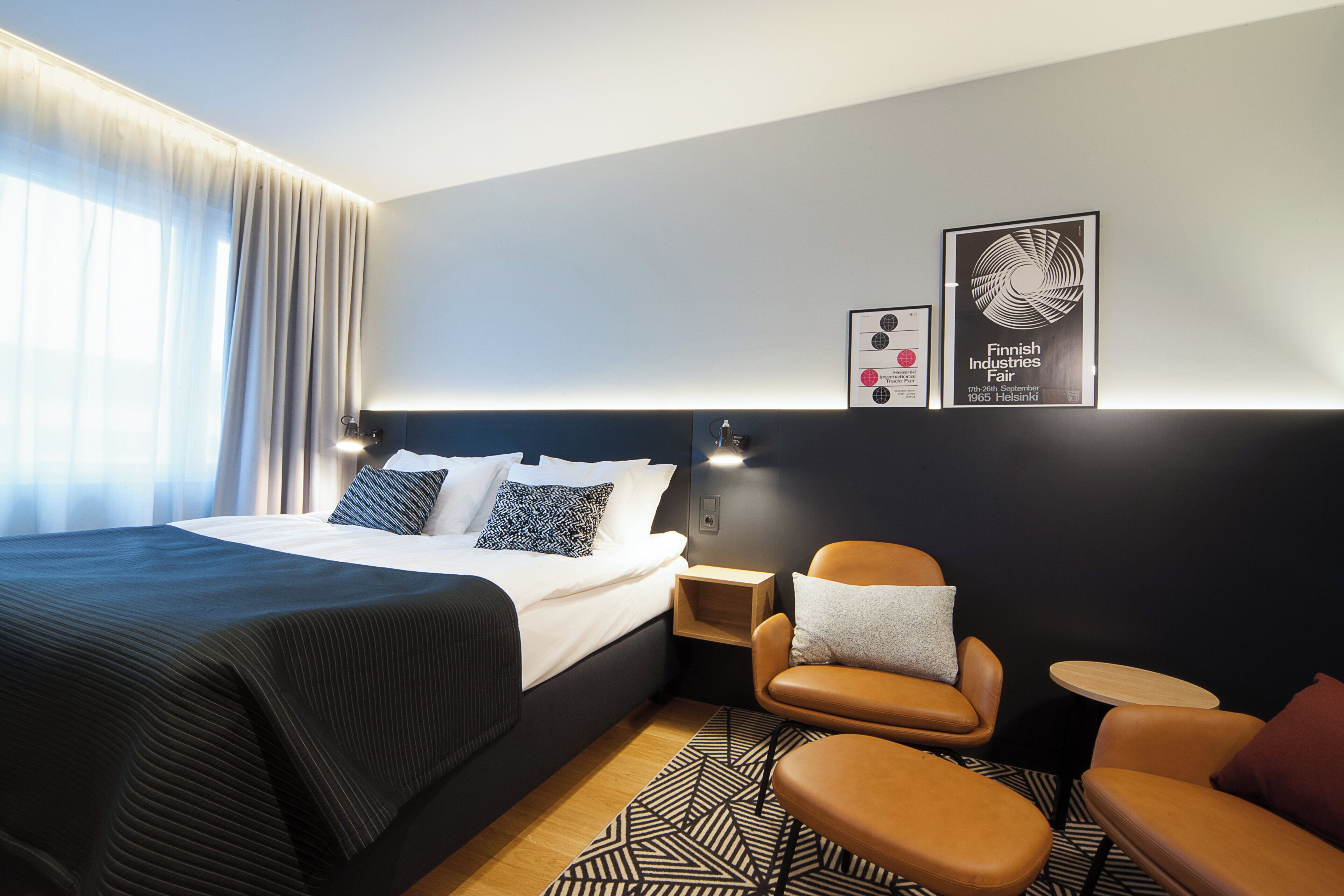 A hotel room with a bed, a chair, a small table, and two framed pictures on the wall.