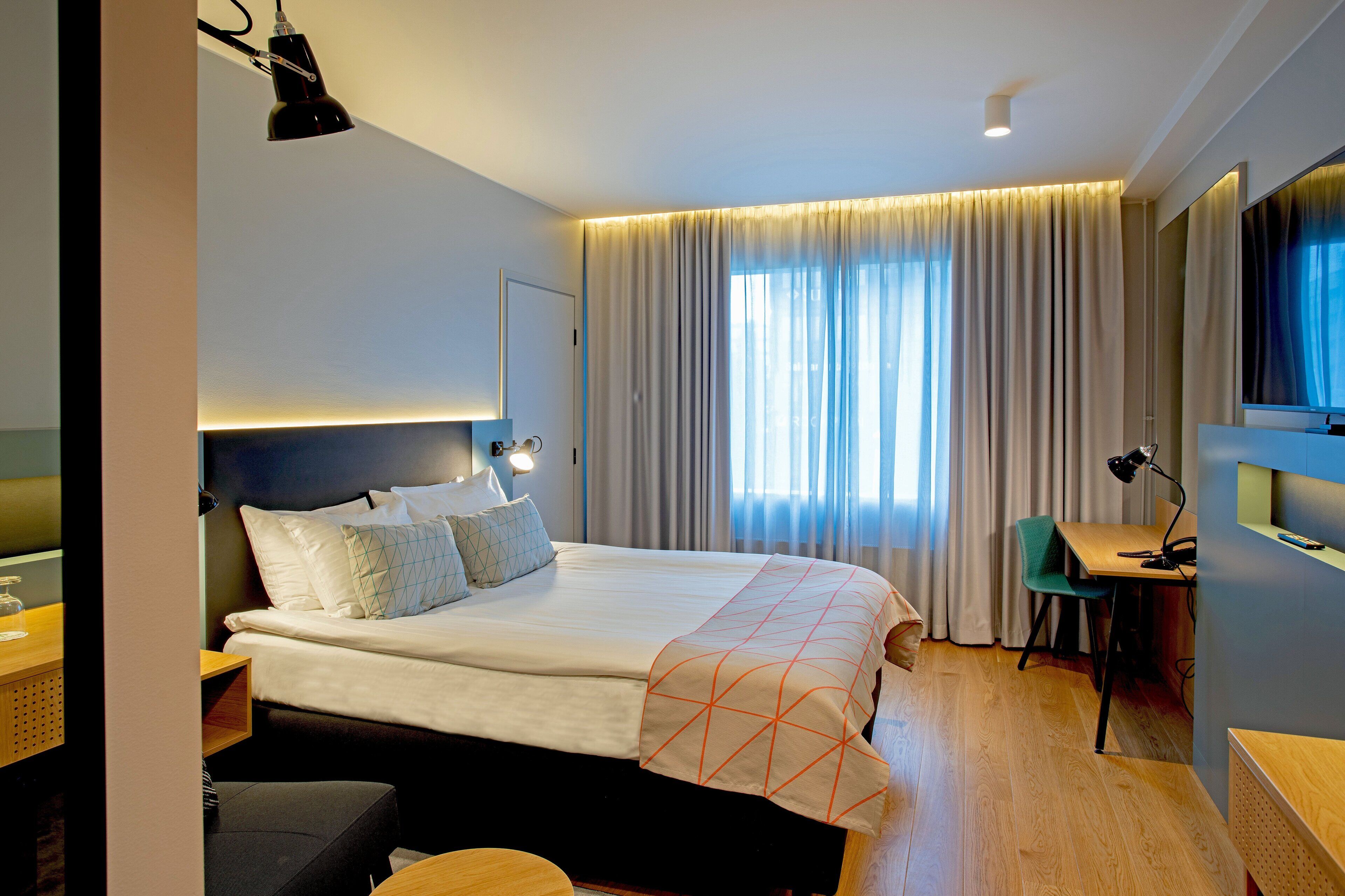 A modern hotel room with a large bed, a desk, a chair, and a TV.