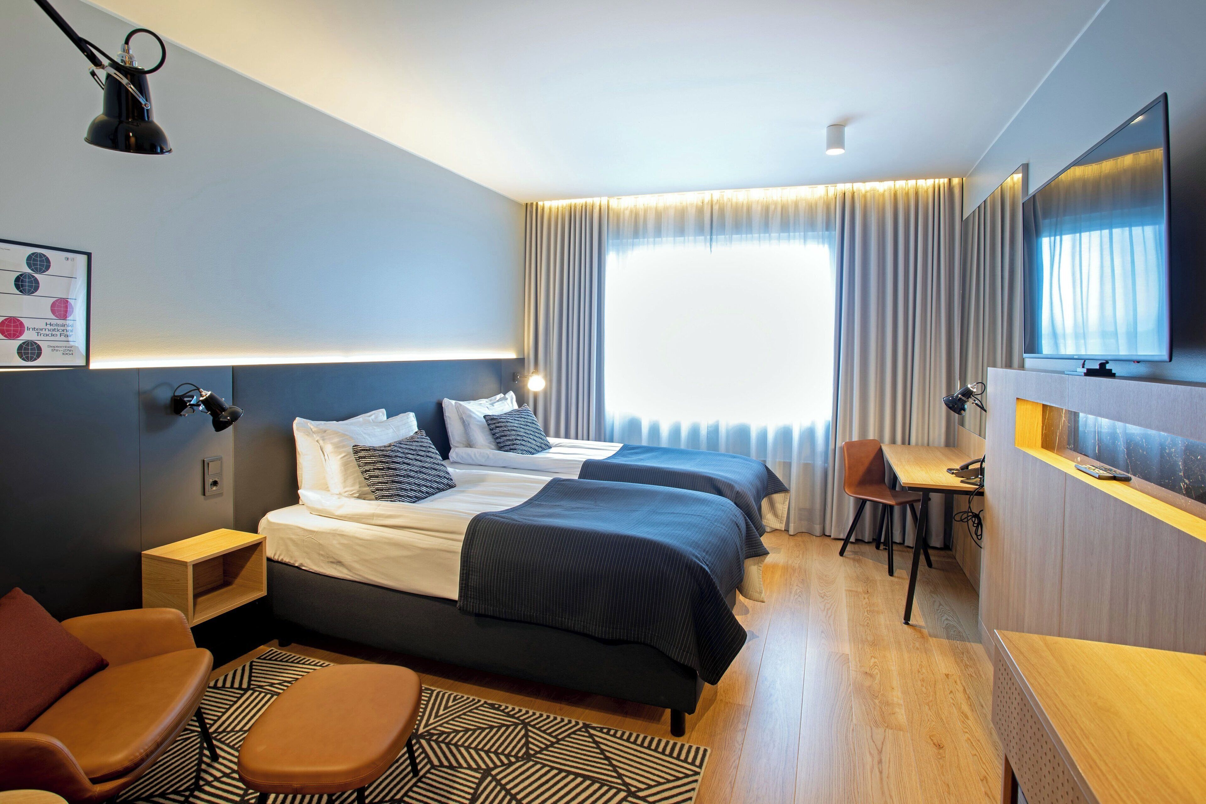 Habitación de hotel con cama, dos mesitas de noche, un escritorio, una silla, un televisor y una ventana con cortinas.