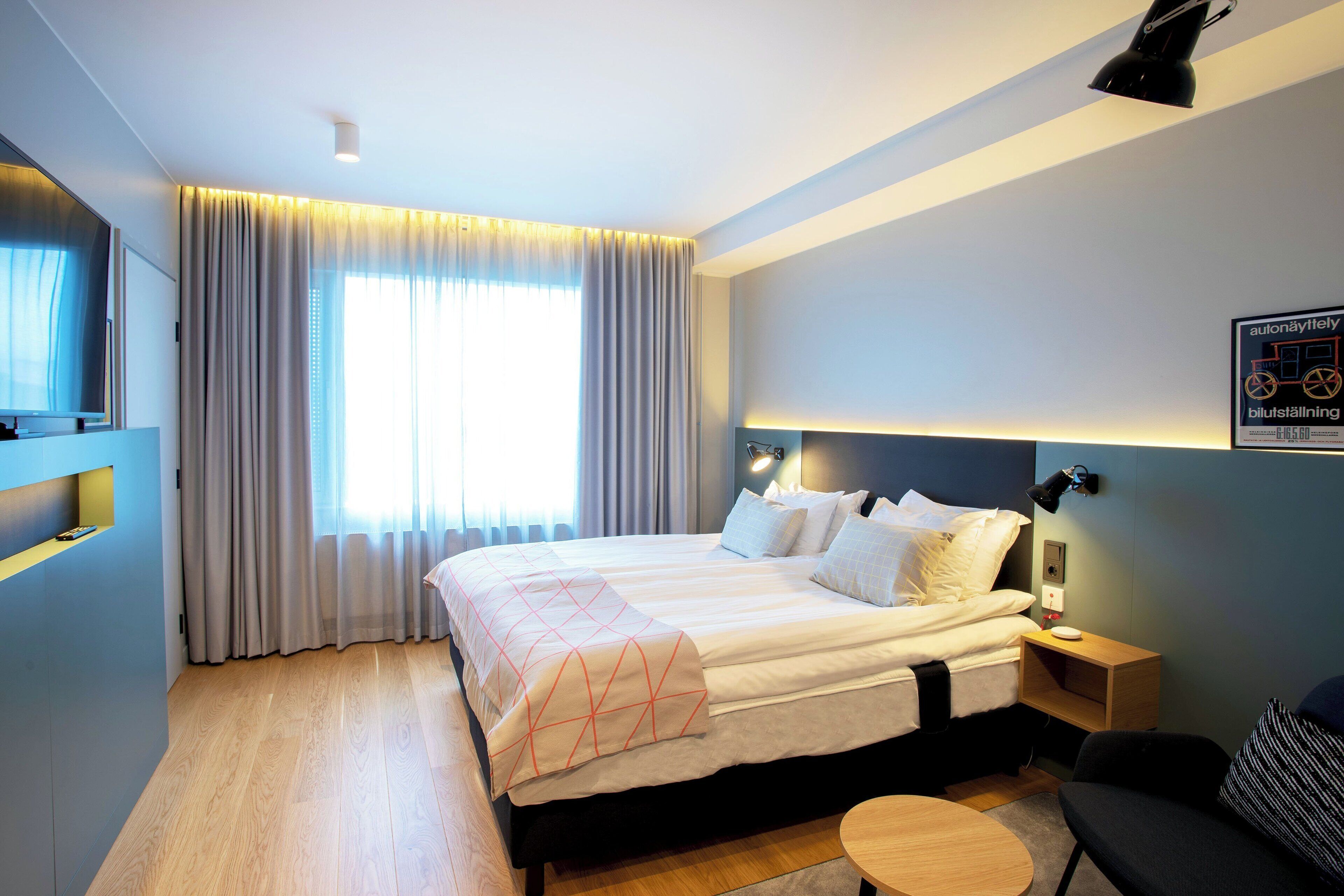 A hotel room with a bed, bedside tables, a TV, and a window with curtains.