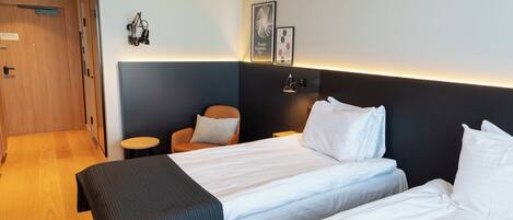 A hotel room with two beds, a small round table, a chair, and a wall-mounted lamp.