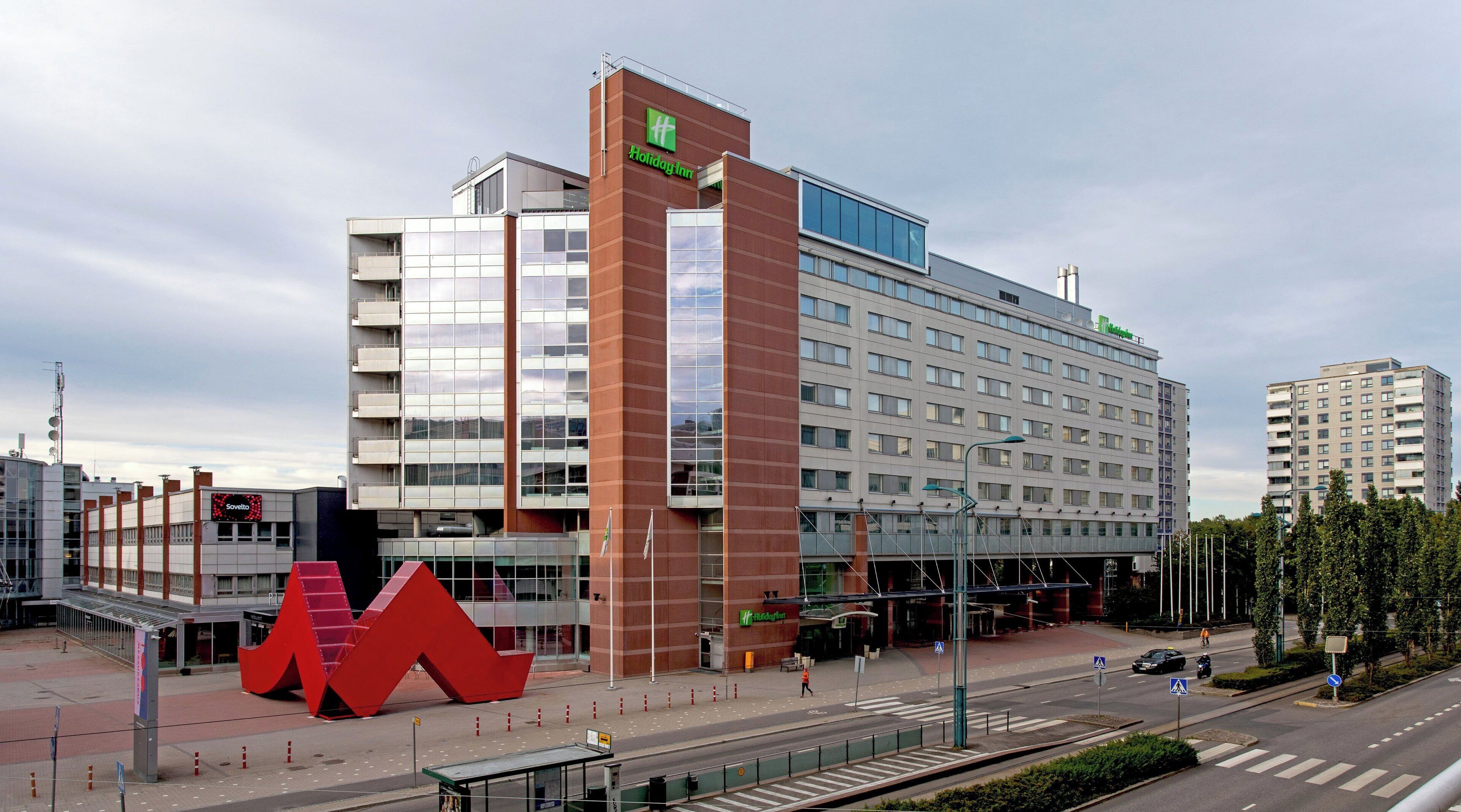 A multi-story hotel with a prominent red 'M' sculpture in front, a Holiday Inn sign, and a visible 'Soviet' sign on a nearby building.