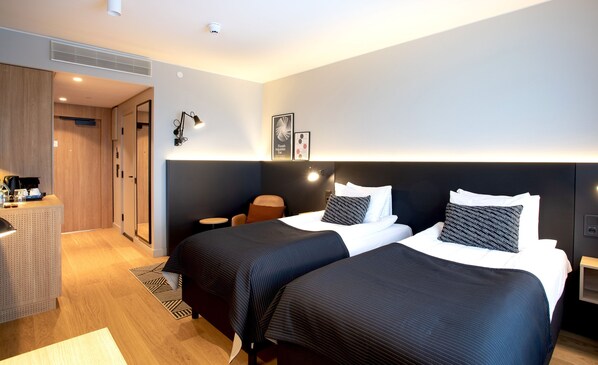 Moderni hotellihuone, jossa on kaksi sänkyä, työpöytä tuolilla ja pieni pöytä.