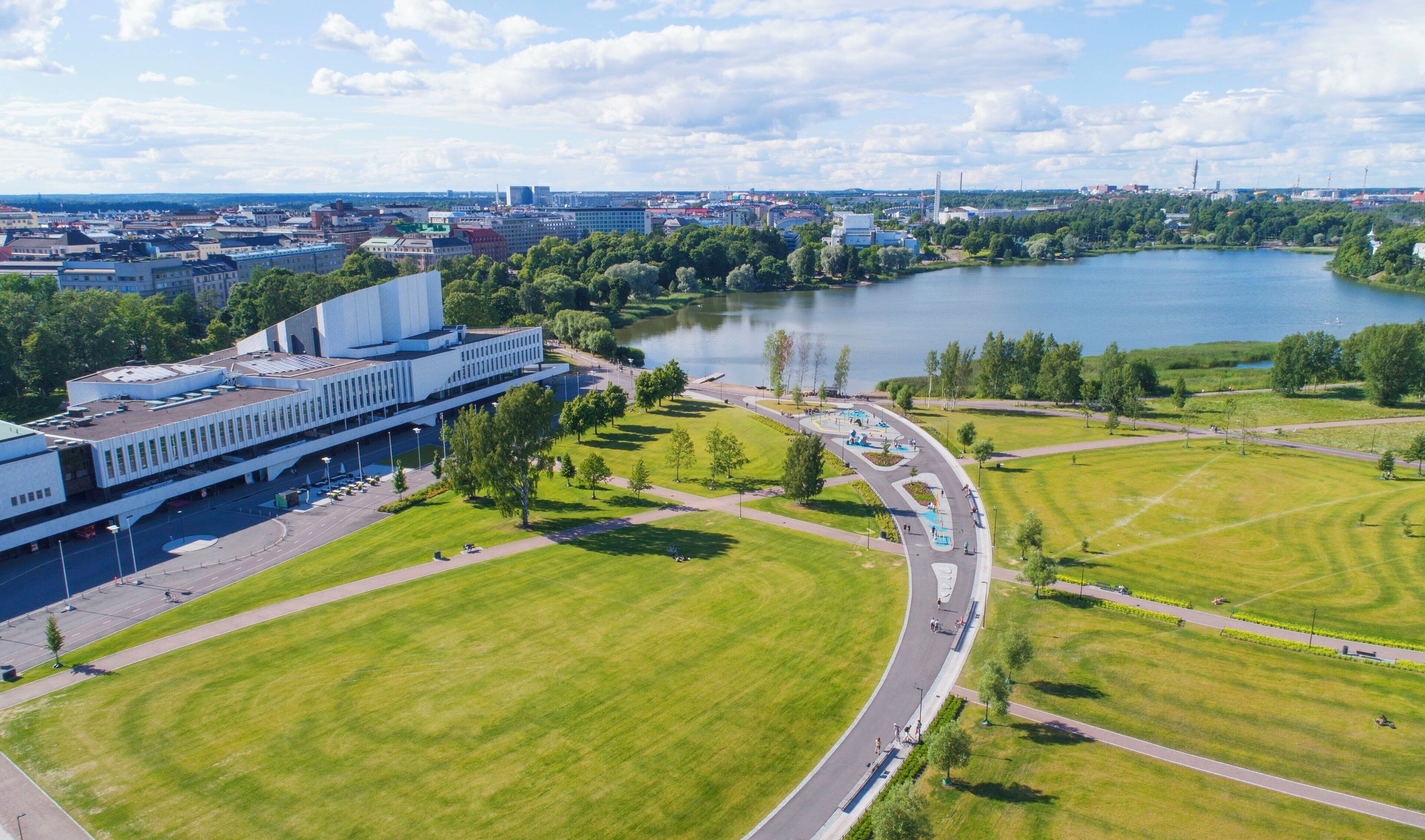 An aerial view of a modern building complex with a large parking area, a lake, and surrounding greenery.