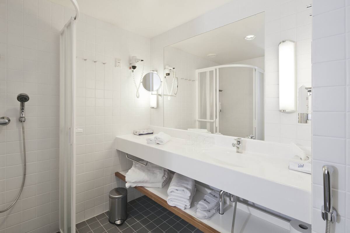 A modern bathroom with a white sink, mirror, and shower. There are towels and toiletries on the counter.
