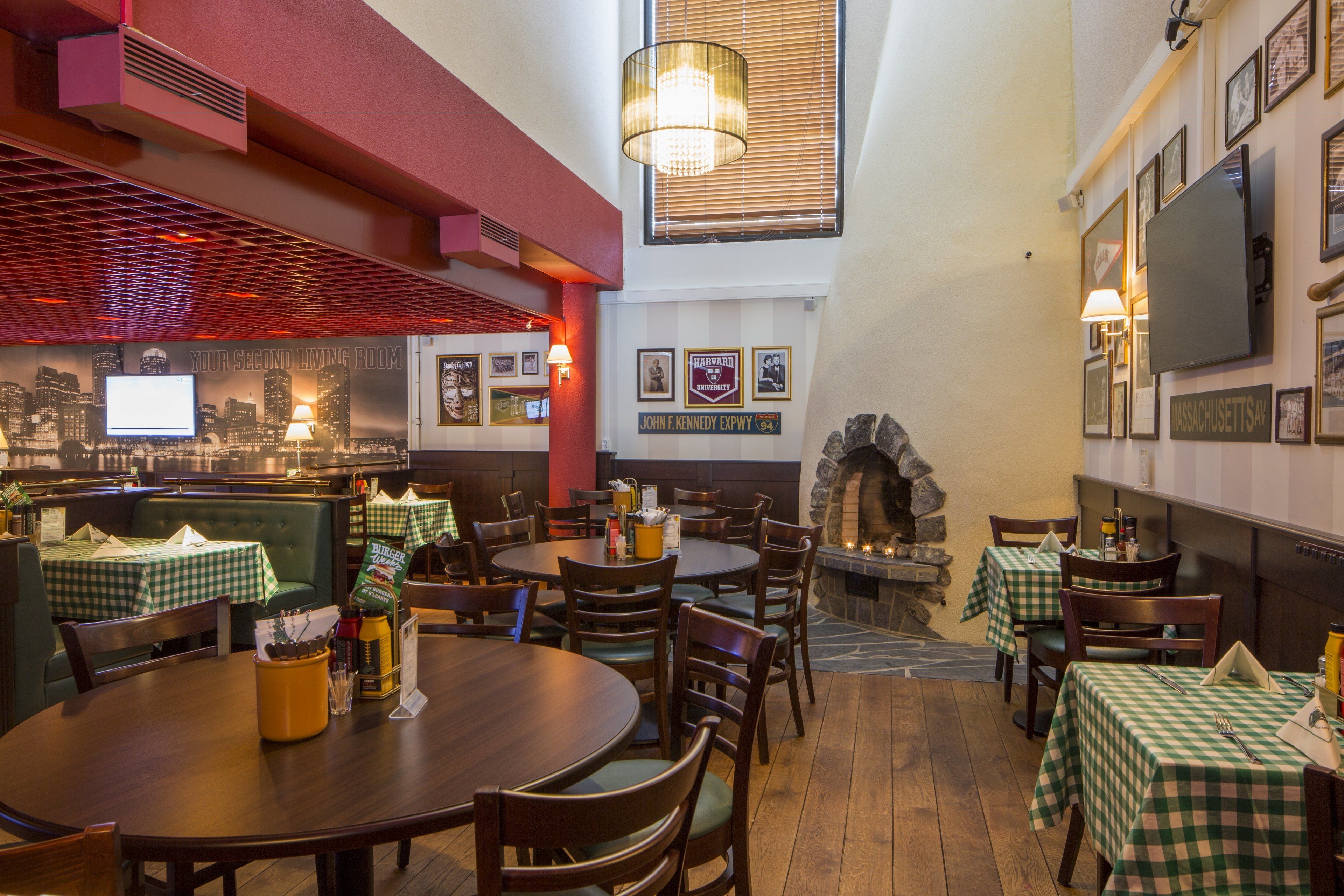 Een restaurantinterieur met ronde tafels, groene geruit tafelkleden, houten stoelen, een stenen openhaard en inlijste foto's aan de wanden.