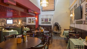 Een restaurantinterieur met ronde tafels, groene geruit tafelkleden, houten stoelen, een stenen openhaard en inlijste foto's aan de wanden.