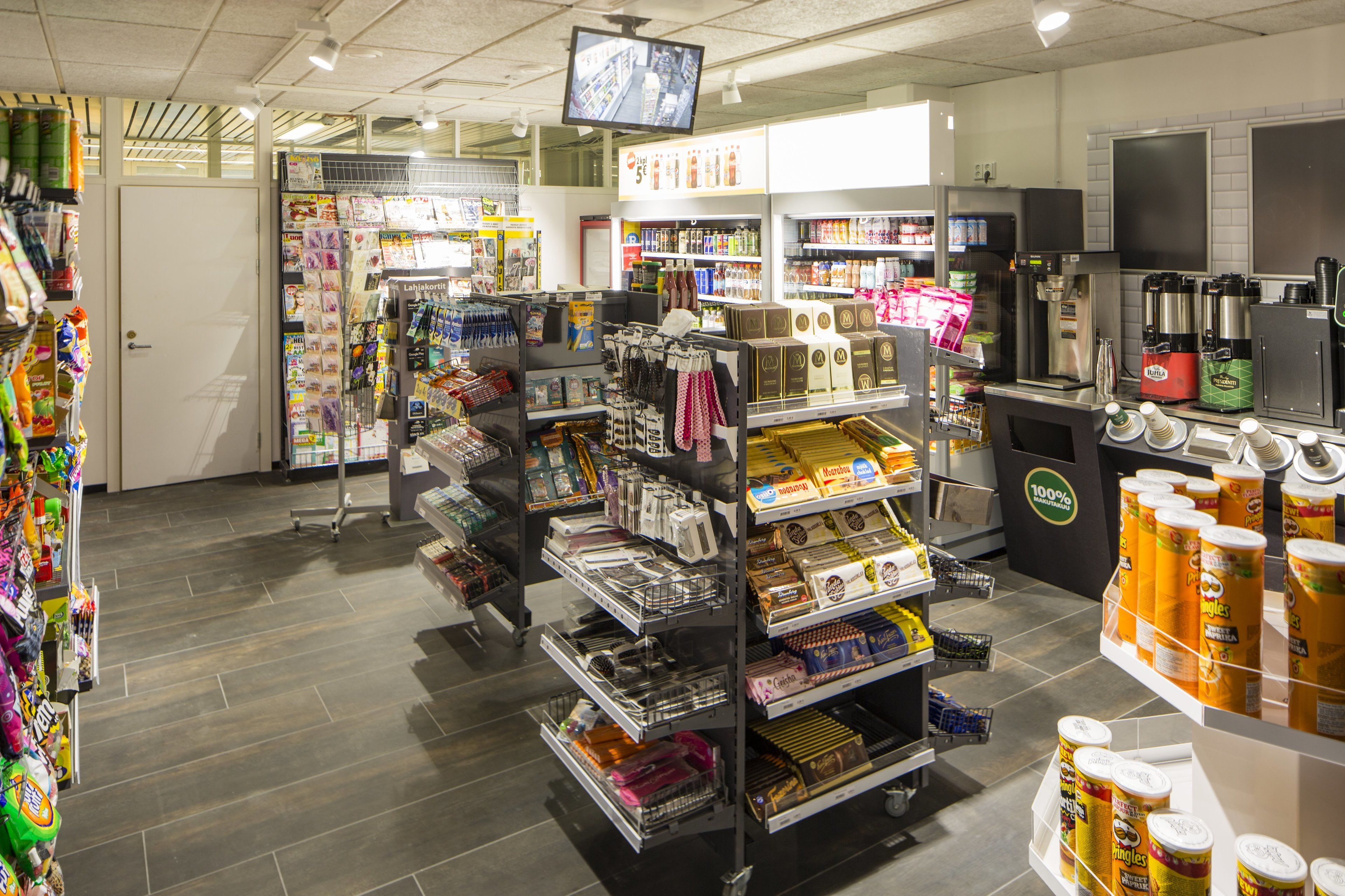 A grocery store interior with shelves stocked with various products, a coffee machine, and a television screen.