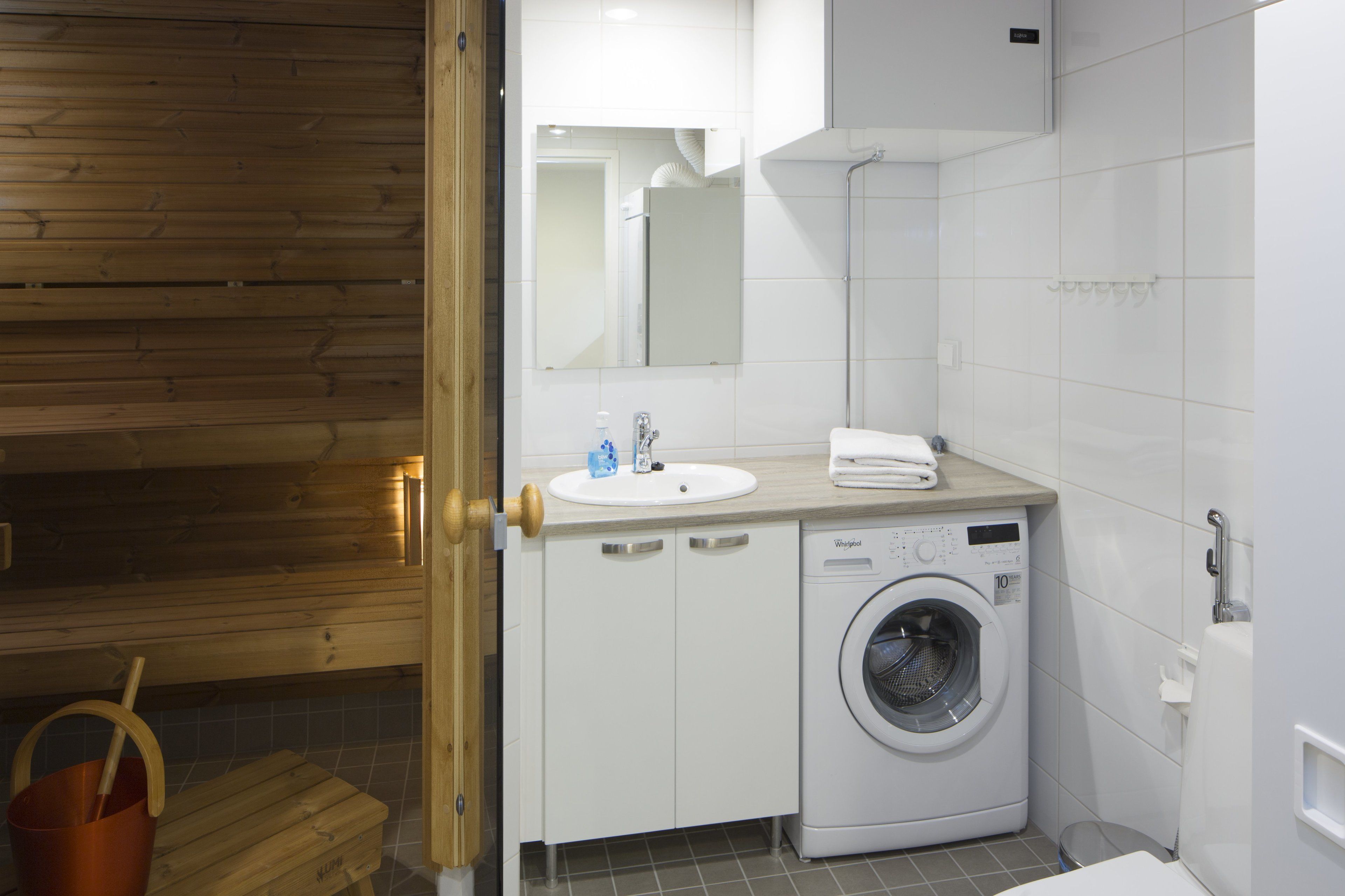 A modern bathroom with a wooden sauna, a white sink, a washing machine, and a tiled floor.