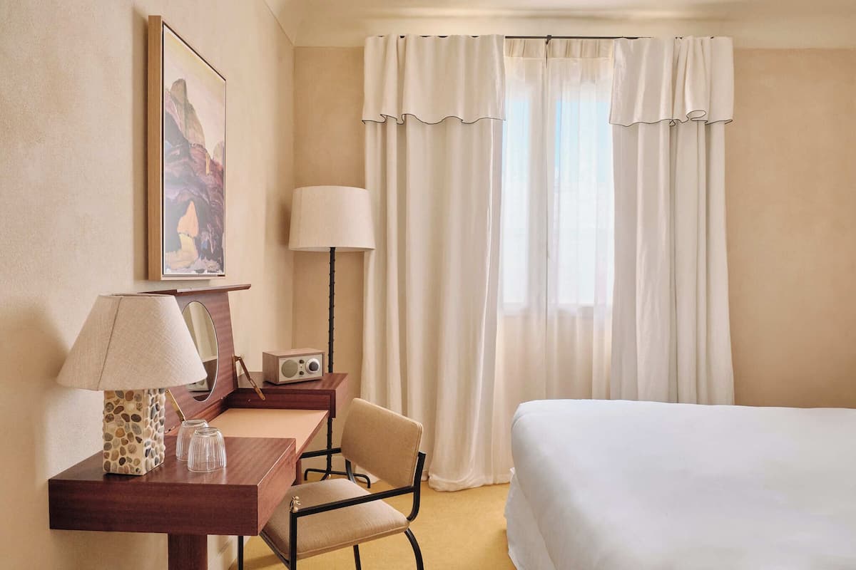 Habitación de hotel con cama, escritorio con lámpara, silla, radio y un cuadro en la pared.