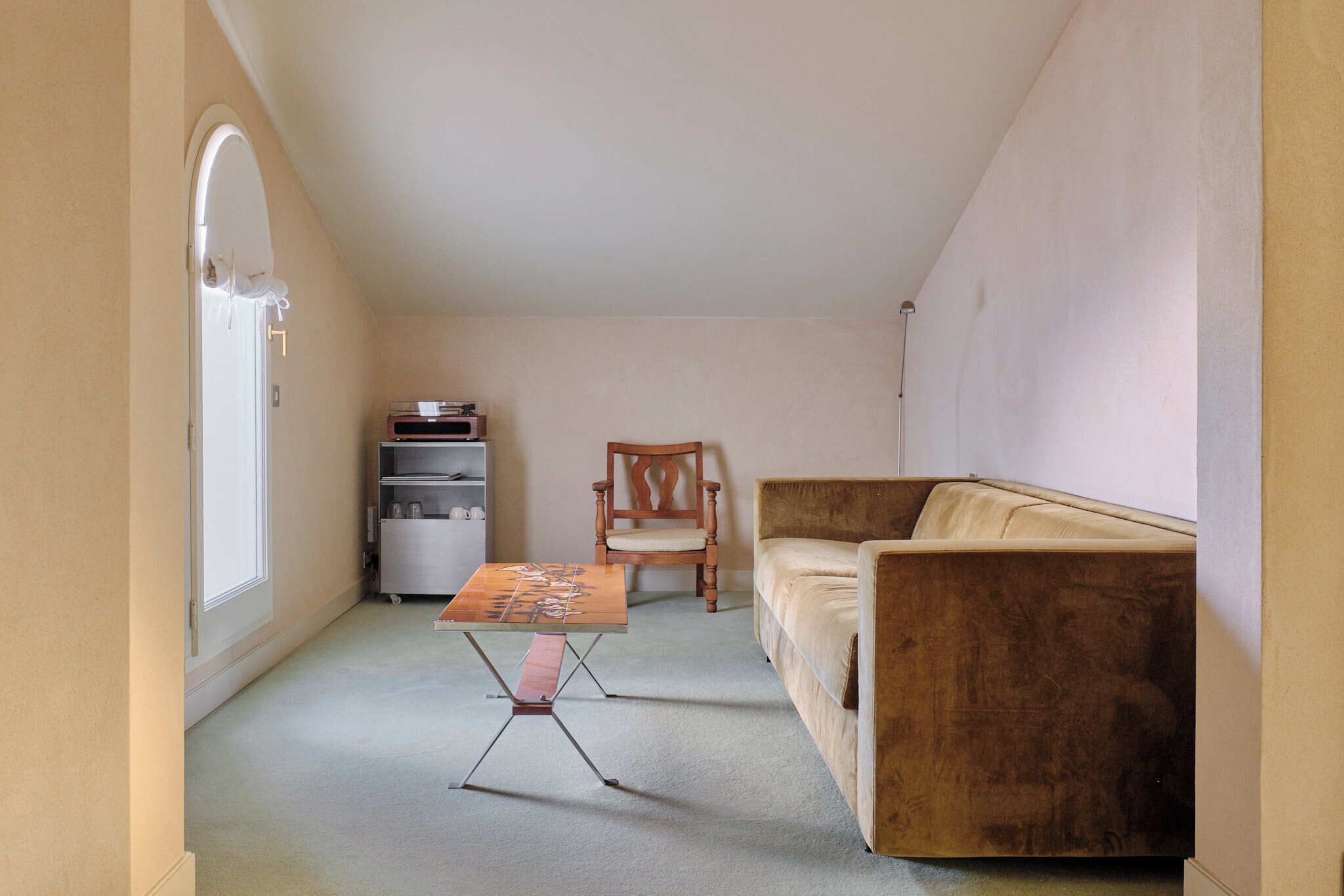 Una habitación pequeña y compacta con un sofá color beige, una silla de madera, una mesita y una puerta blanca.