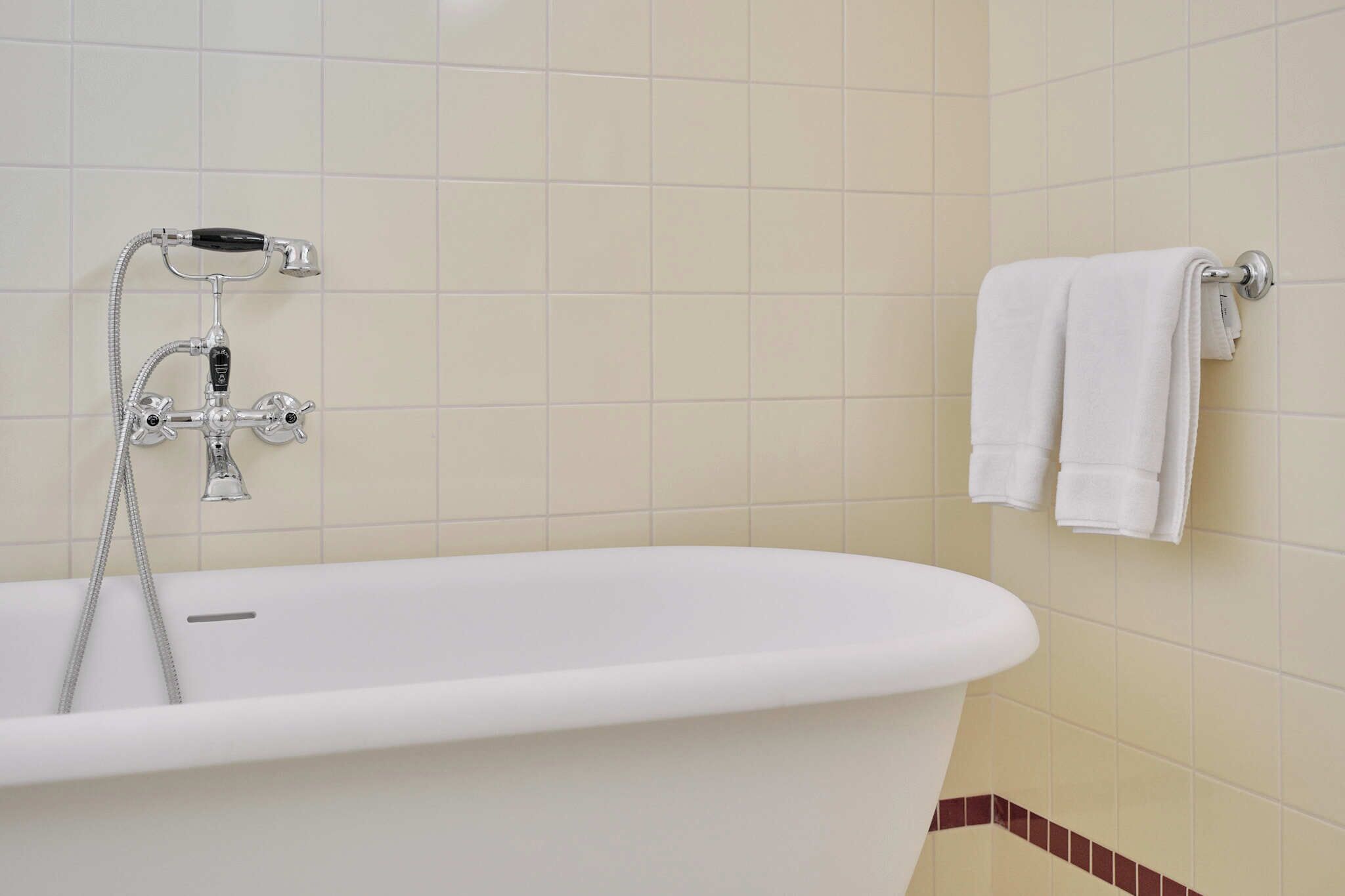 Una bañera blanca con un cabezal de ducha manual y un grifo instalado en la pared. Hay dos toallas blancas colgadas en un perchero.