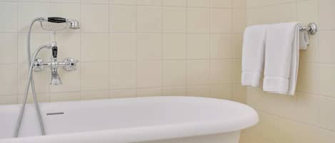 Una bañera blanca con un cabezal de ducha manual y un grifo instalado en la pared. Hay dos toallas blancas colgadas en un perchero.
