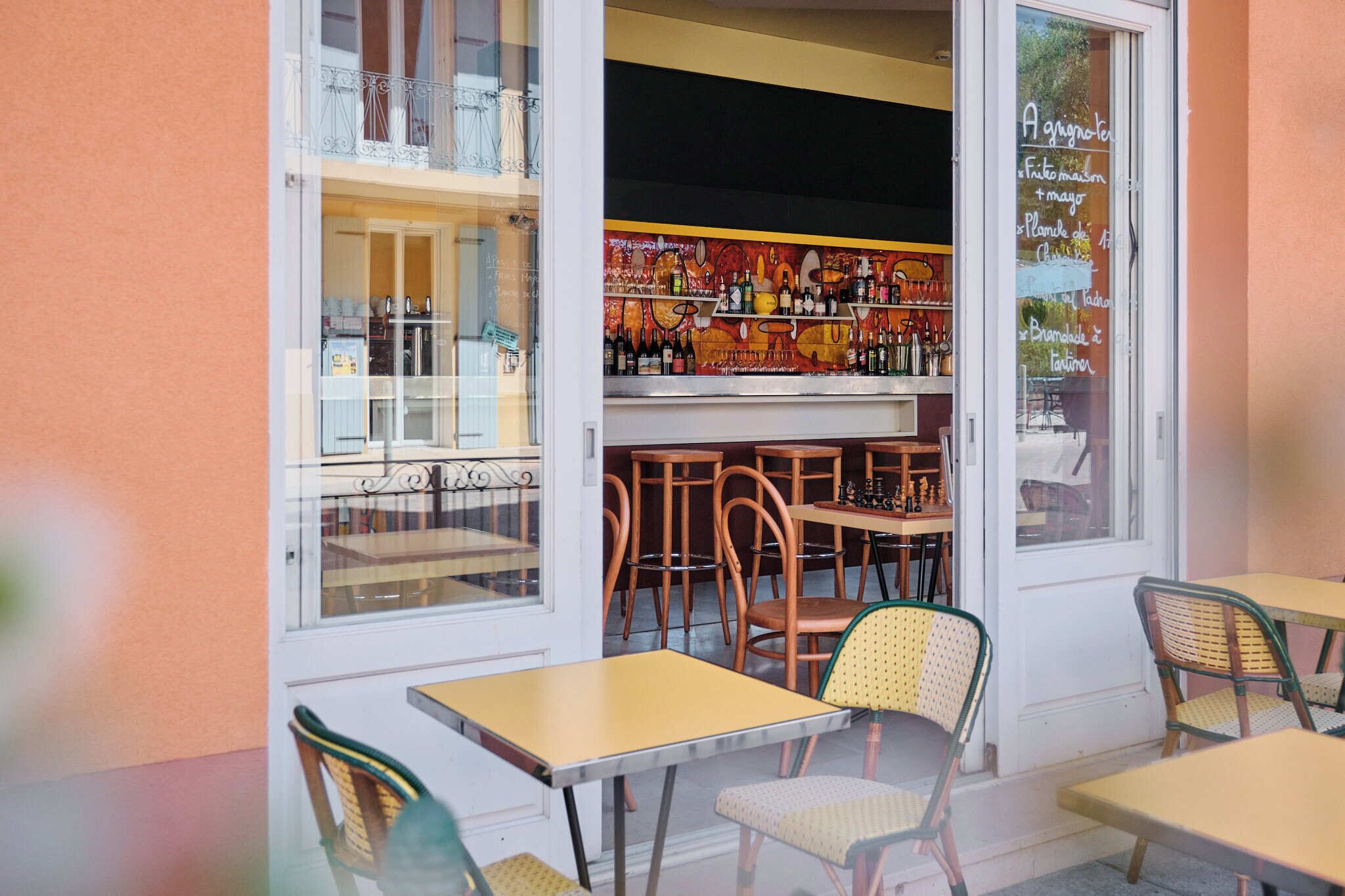 An outdoor seating area with tables and chairs, a bar with shelves of bottles, and a view of the street through large windows.