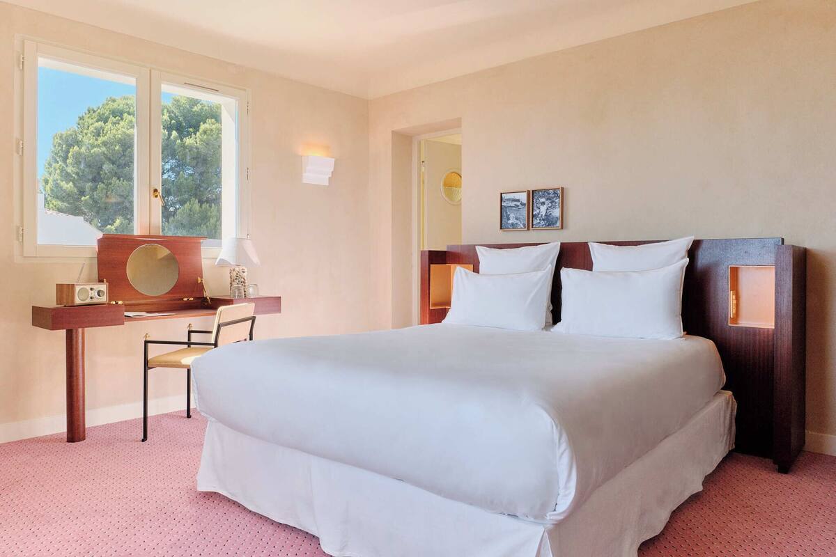 Una habitación de hotel con una cama grande, un escritorio con una silla, un espejo redondo y dos cuadros en la pared.