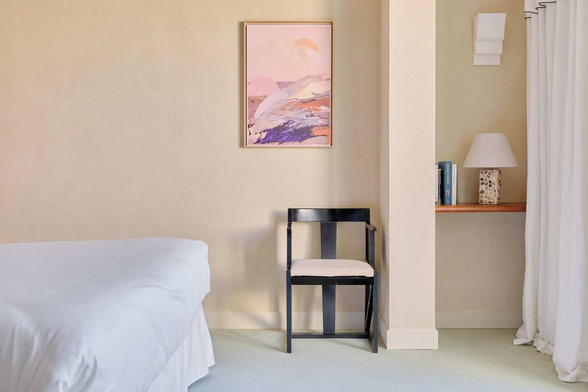 Una habitación de hotel con una cama, una silla, un cuadro en la pared, una lámpara y una estantería con libros y un objeto decorativo.