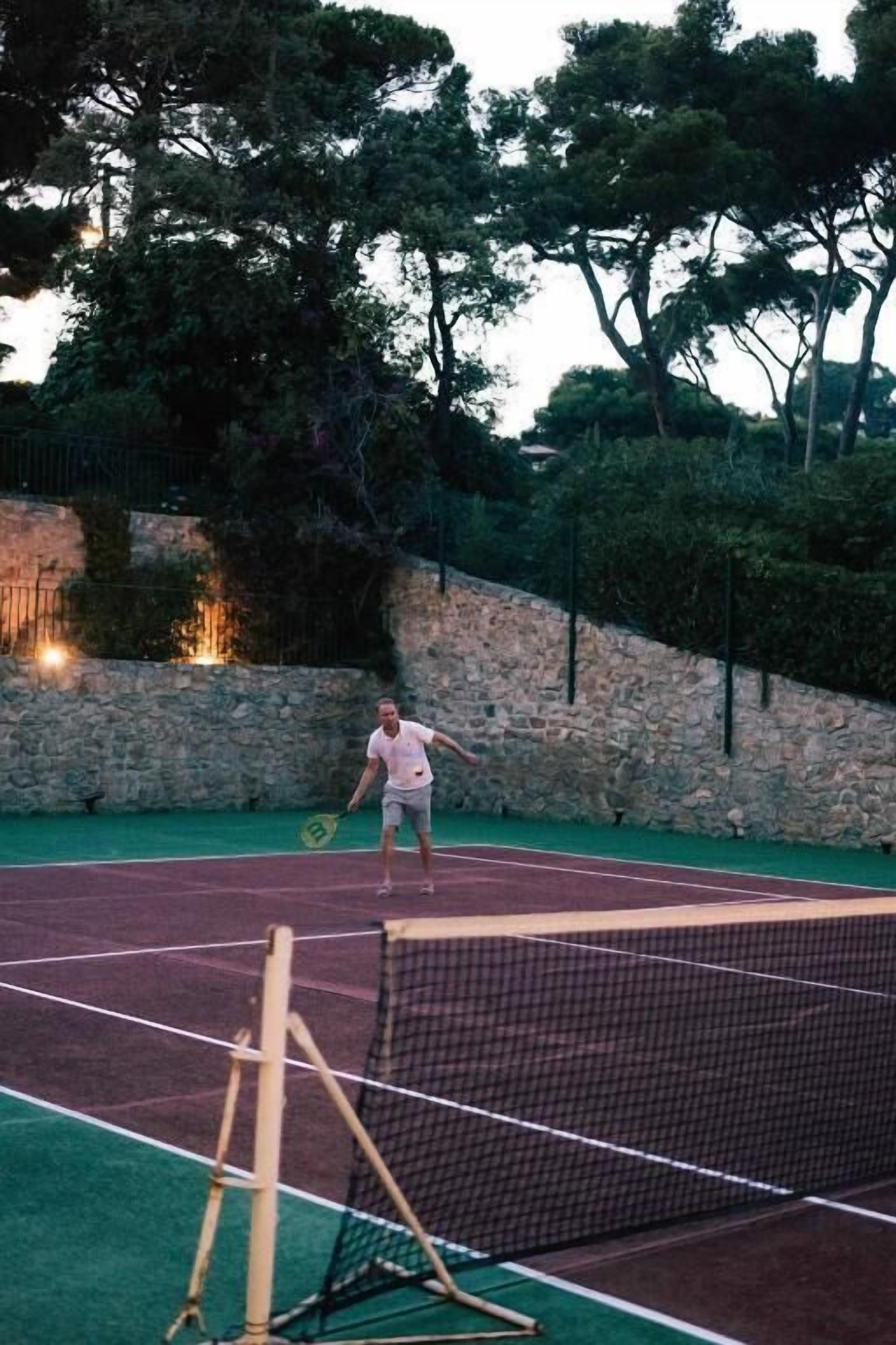 A tennis court with a net, surrounded by a stone wall and trees.