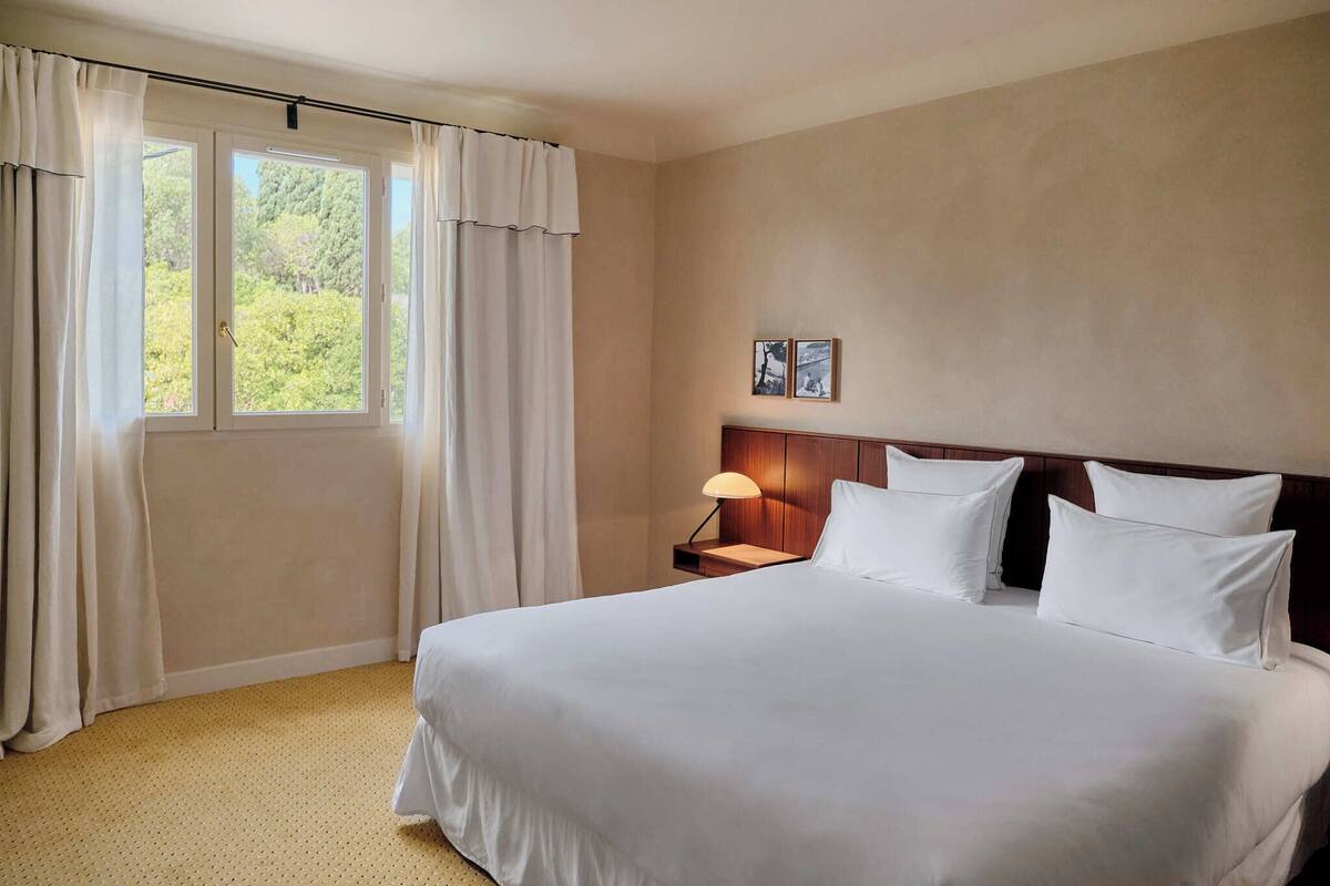 Una habitación de hotel con una cama grande, una mesita de noche con una lámpara, una ventana con cortinas y un cuadro en la pared.