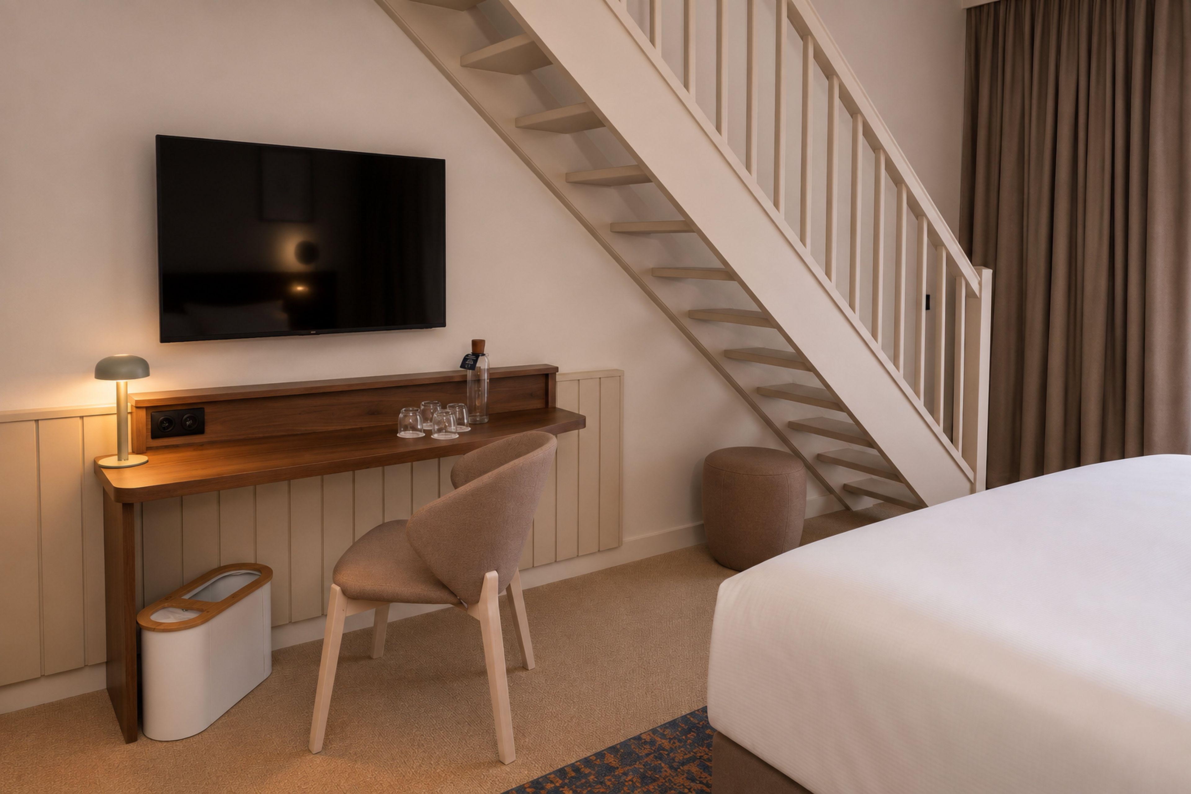 Ein Hotelzimmer mit einem Bett, einem Schreibtisch mit Fernseher, einem Sessel, einem Papierkorb und einer Treppe.