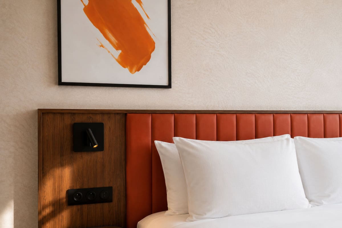 Ein Hotelzimmer mit Nachttisch aus Holz, einem an der Wand befestigten Lautsprecher, einem roten, mit Stoff bezogenen Kopfteil und weißer Bettwäsche.