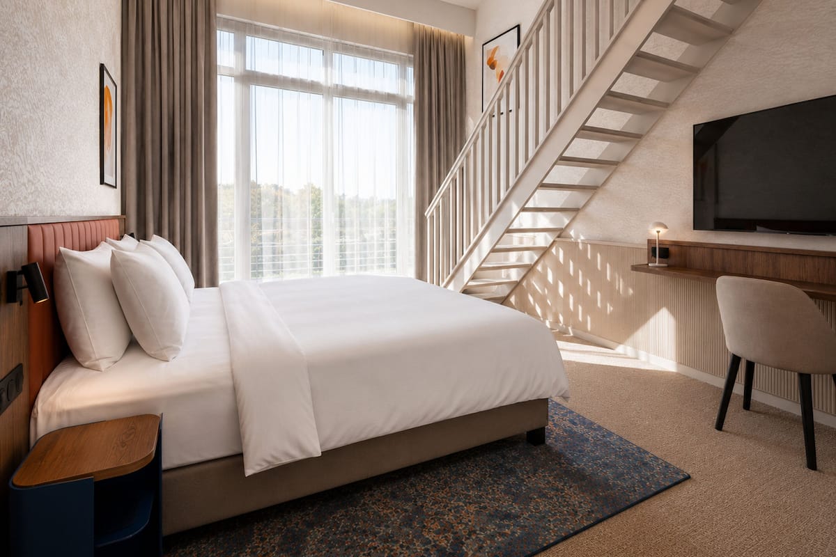 Ein Hotelzimmer mit einem Bett, einem Schreibtisch mit Fernseher, einem Sessel, einer Treppe und einem Fenster mit Vorhängen.