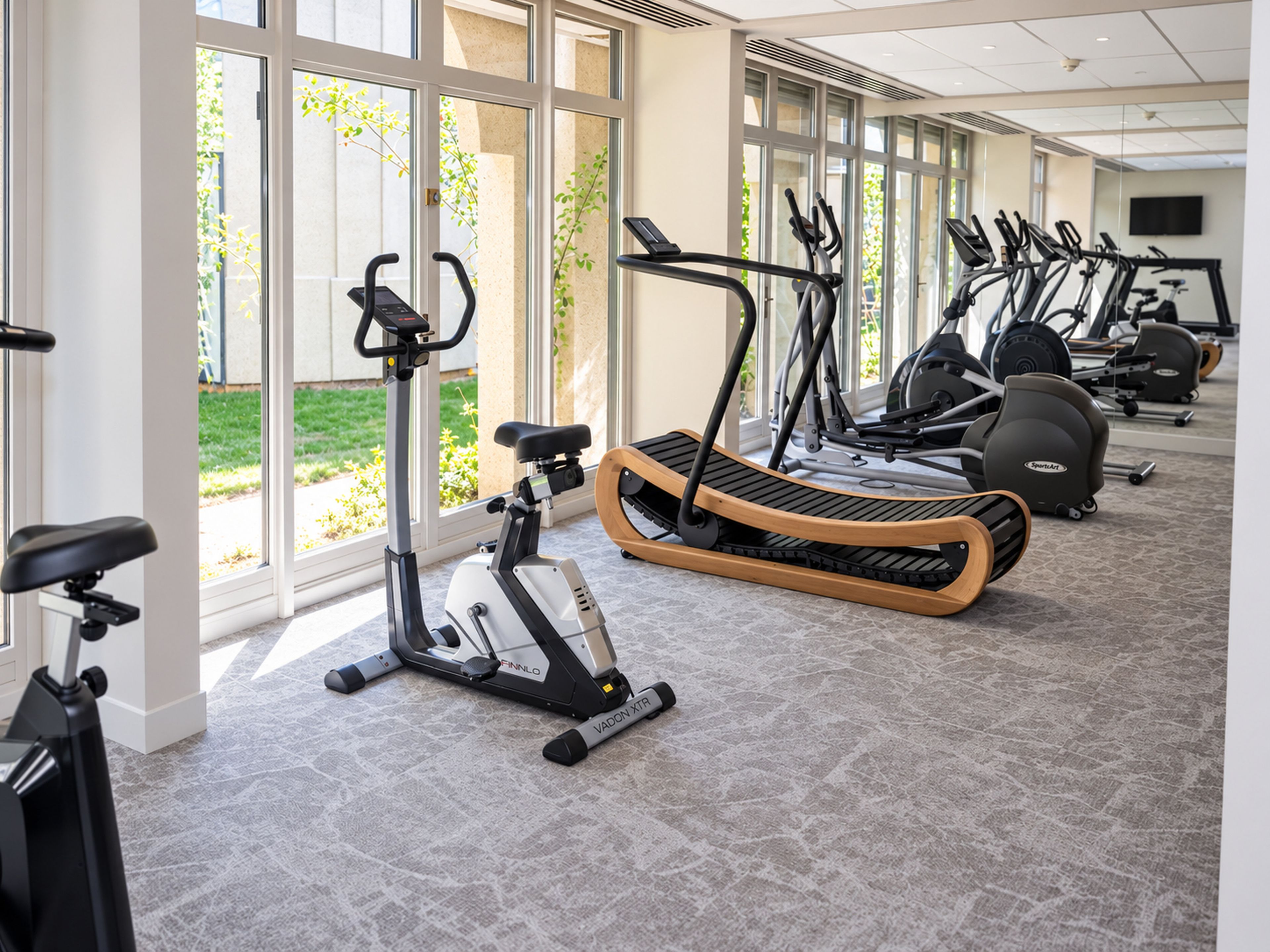 Une salle de sport équipée de divers appareils de cardio, dotée de grandes fenêtres et d’un sol carrelé.