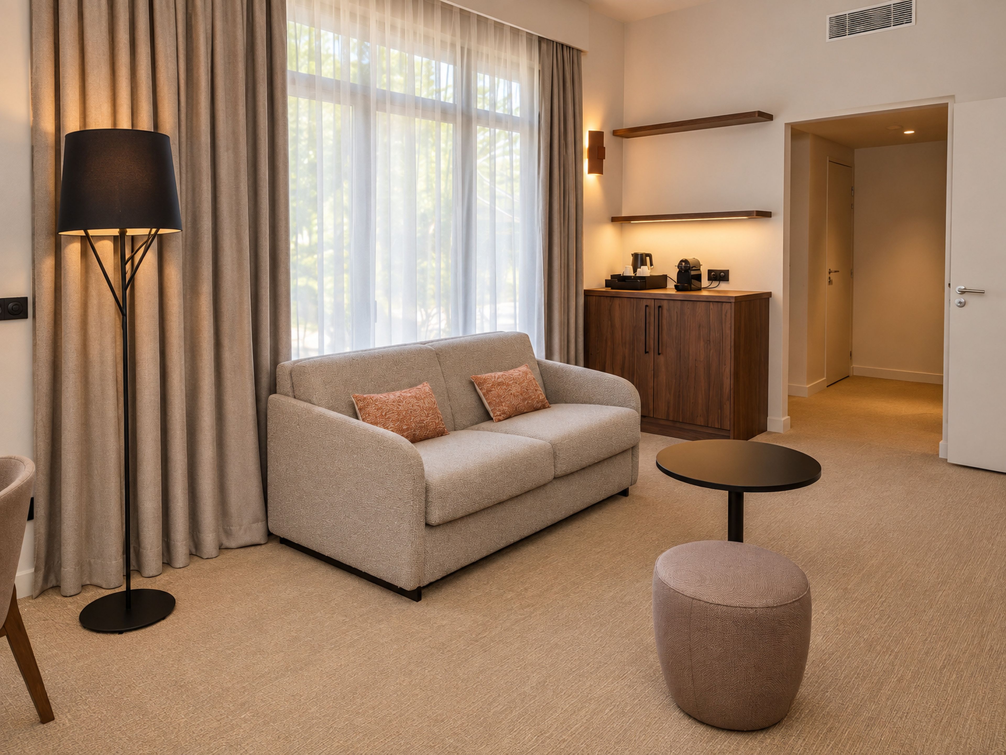 Una habitación de hotel moderna con sofá, reposapiés, lámpara de pie y escritorio con cafetera.