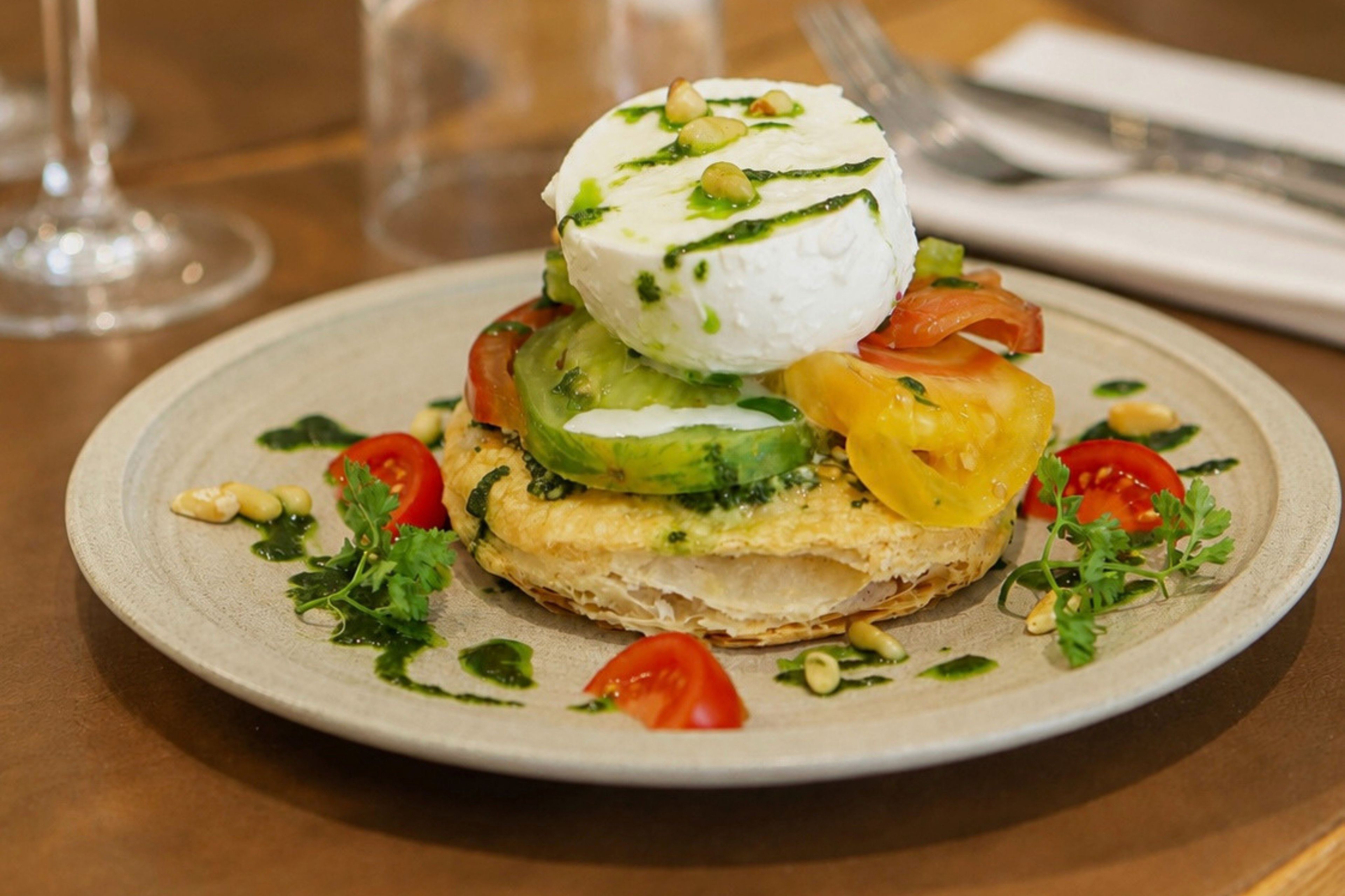 A plate with a layered dish, garnished with herbs and nuts, on a table with a glass and a napkin.