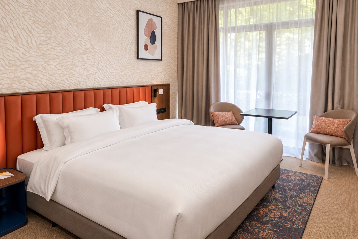 Een hotelkamer met een groot bed, een klein tafeltje, twee stoelen, een hoofdbord en een raam met gordijnen.