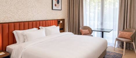 Habitación de hotel con una cama grande, una mesita pequeña, dos sillas, un cabecero y una ventana con cortinas.