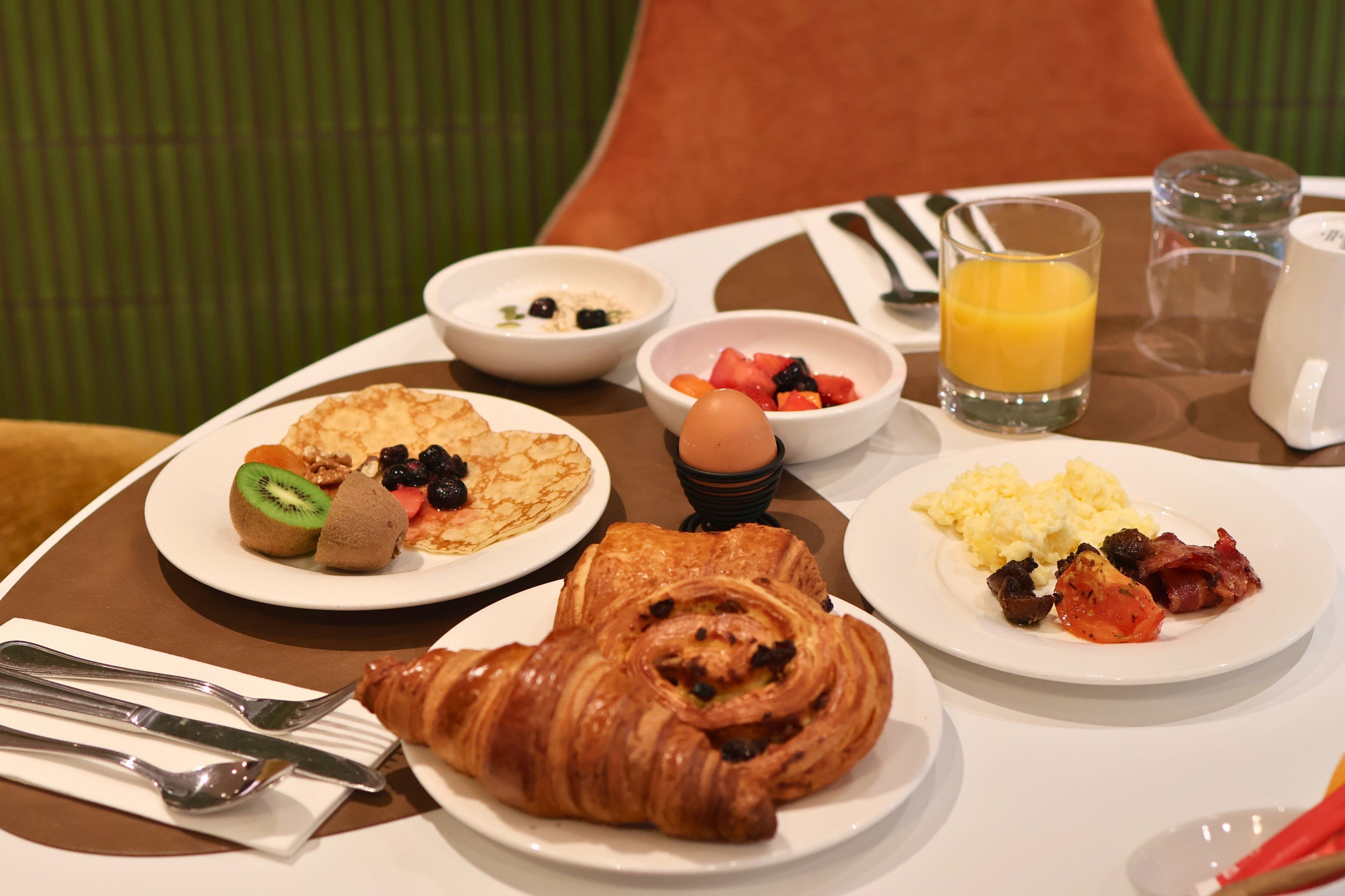 Une table mise pour le petit-déjeuner avec des assiettes de nourriture, un croissant, un verre de jus d’orange et un bol de fruits.