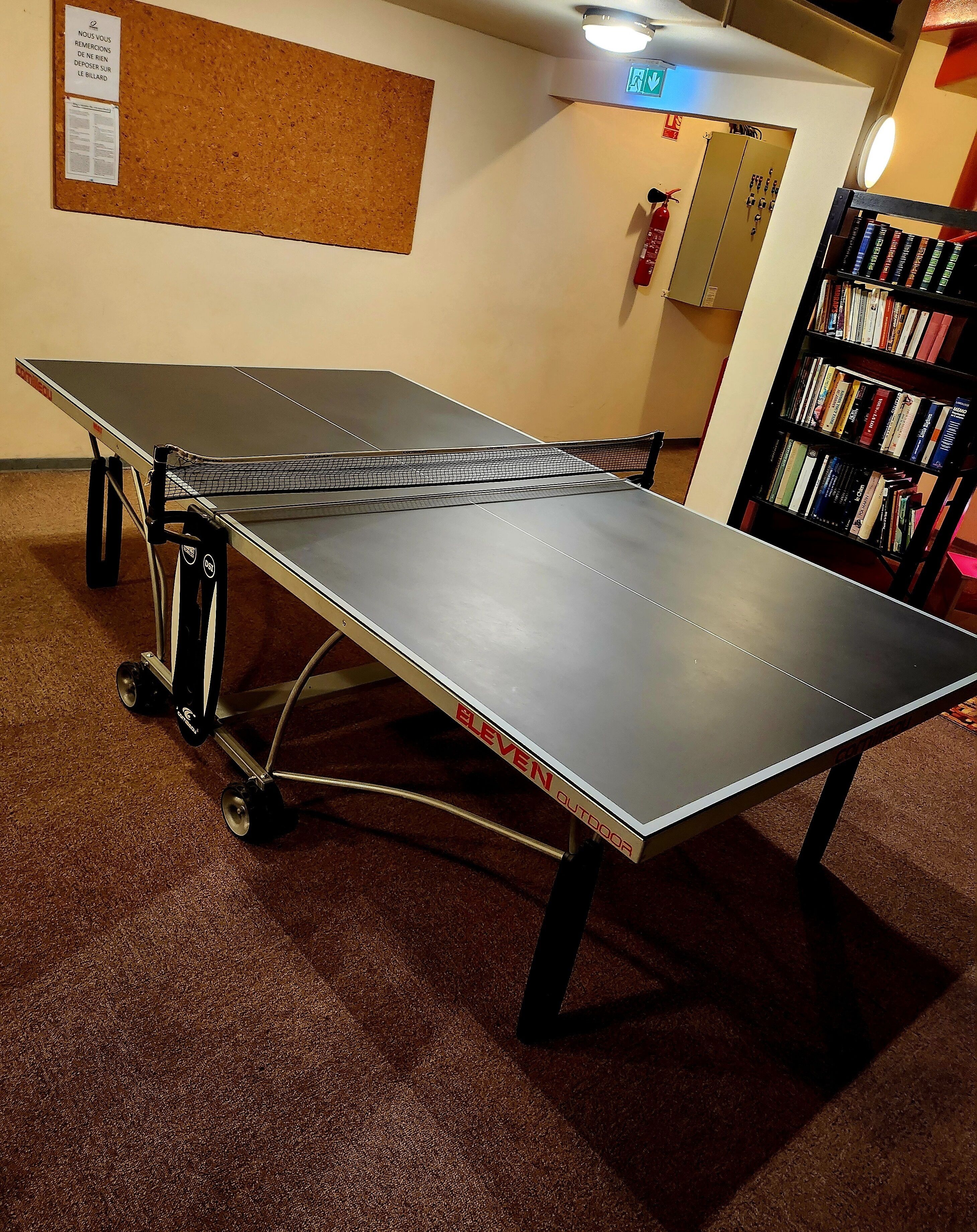 Een tafeltennistafel op wielen, een prikbord met mededelingen, een brandblusser en een boekenkast.