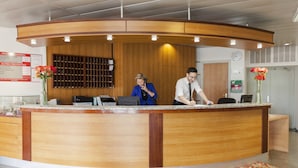 Un comptoir d’accueil avec deux employés, un comptoir en bois, une horloge et un tableau d’affichage où est affichée une liste de prix.