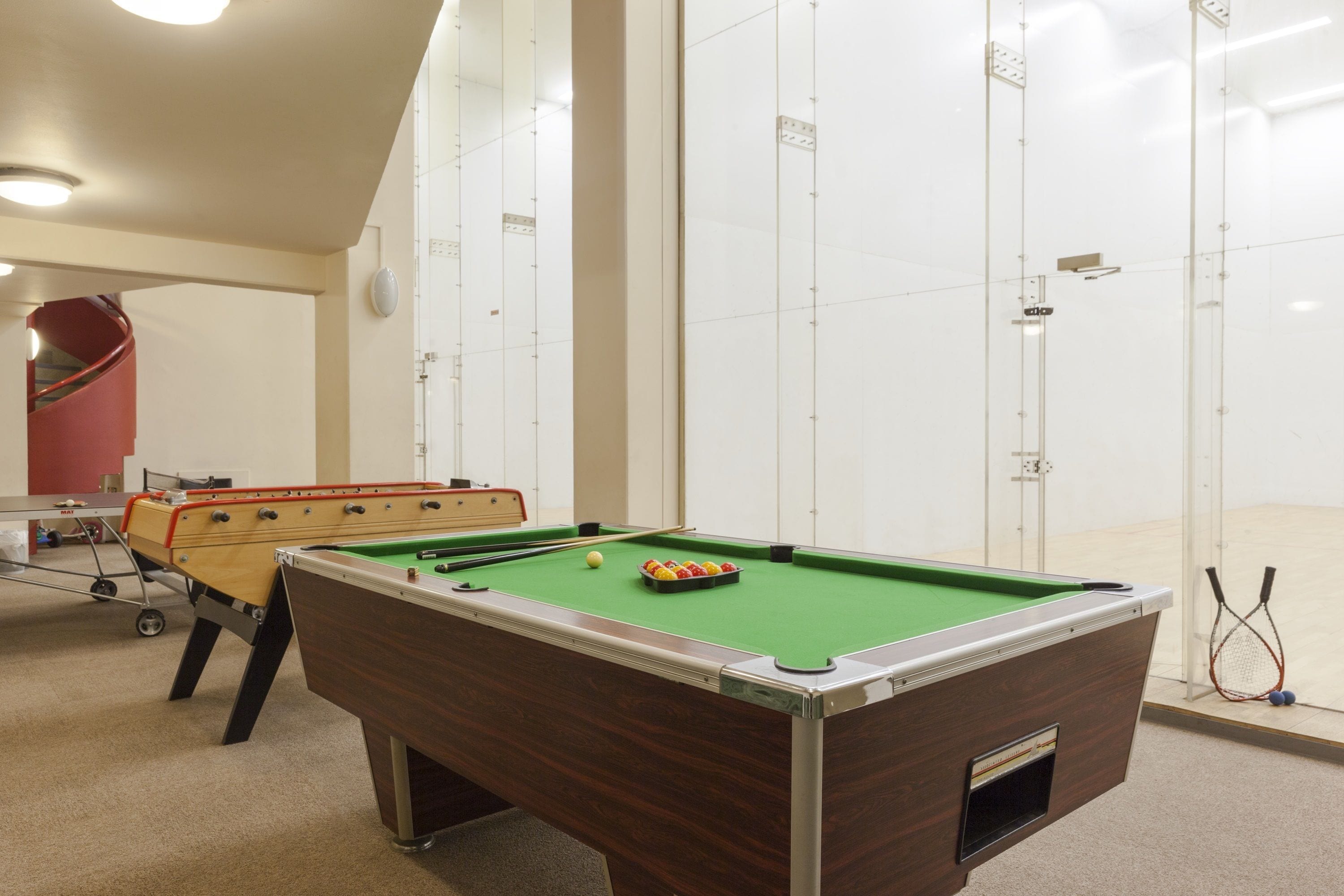 A pool table in a room with a red spiral staircase and a squash court visible through glass doors.