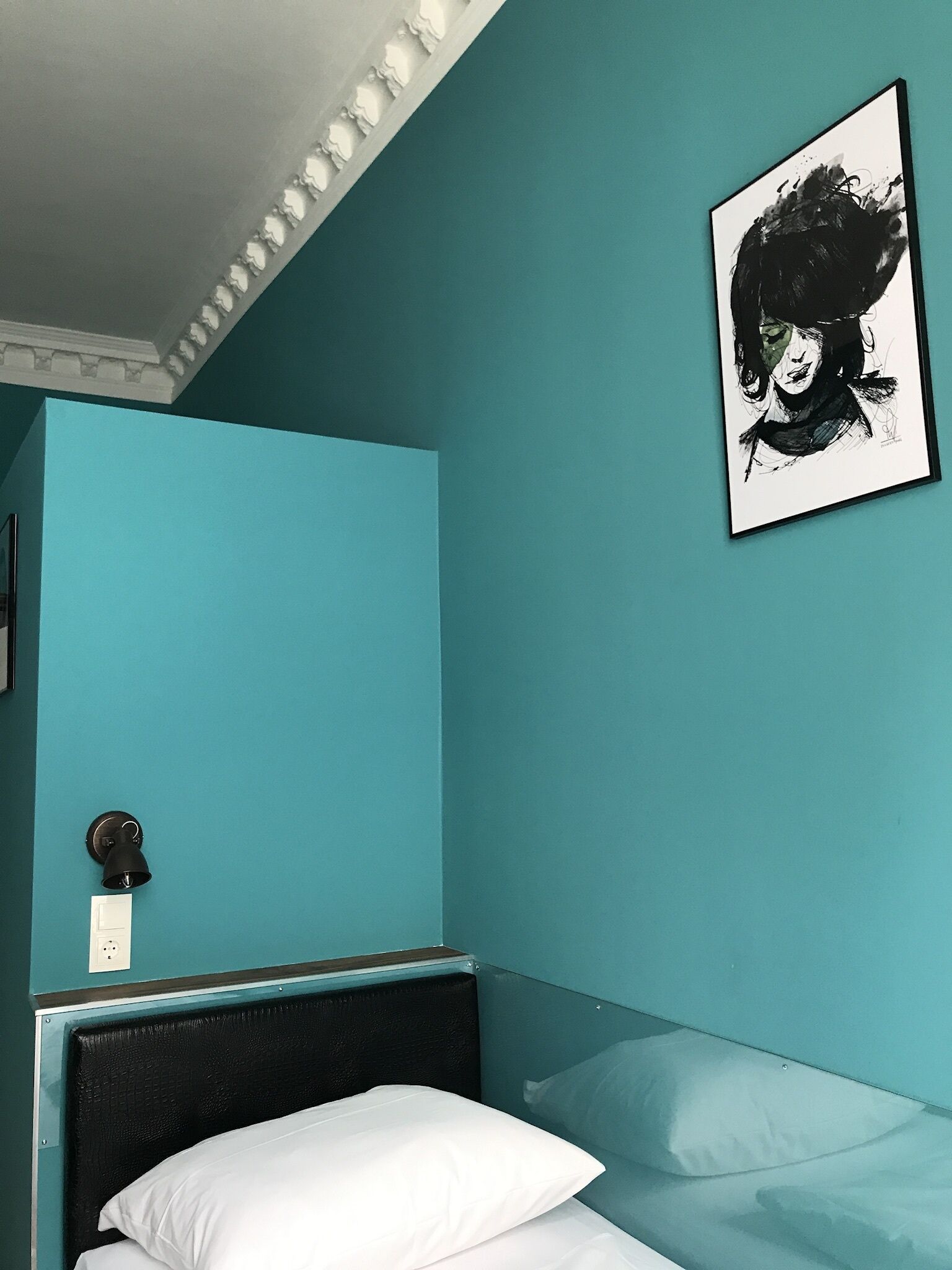 Un dormitorio con paredes color turquesa, cabecera negra, ropa de cama blanca, lámpara negra y un retrato blanco y negro enmarcado.