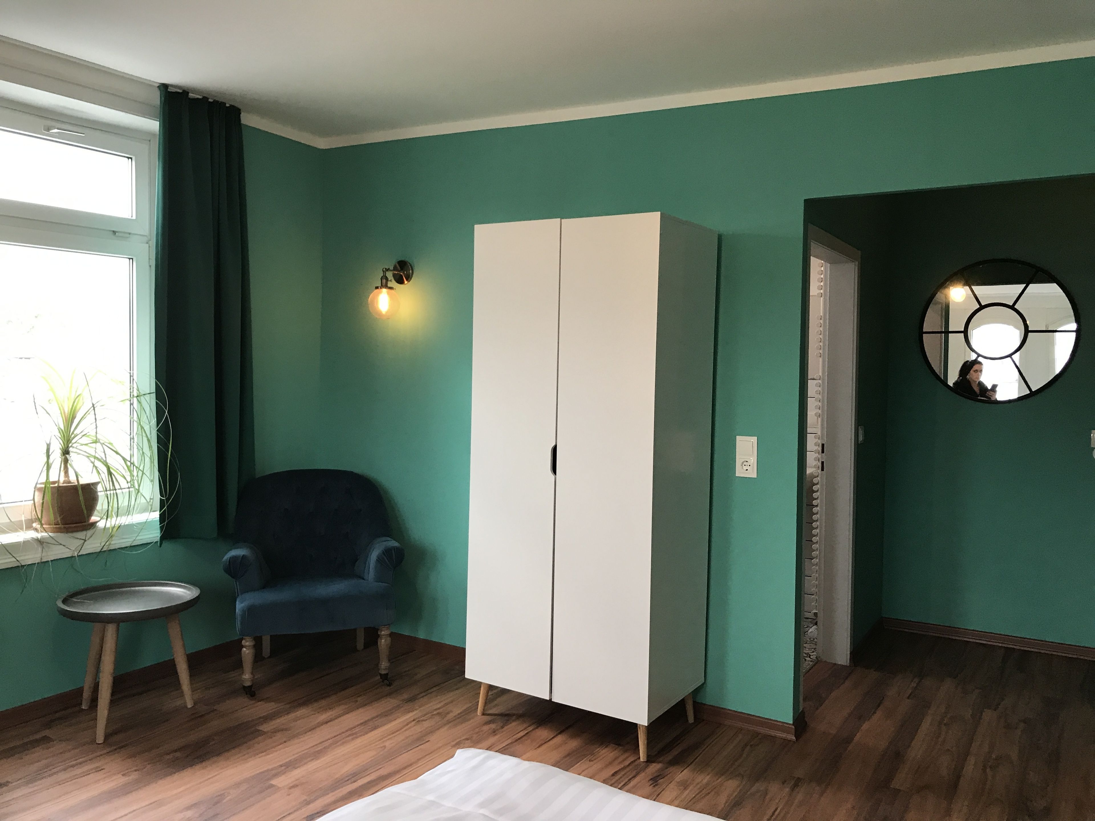 A room with teal walls, a white wardrobe, a blue armchair, a small round table, and a potted plant by the window.