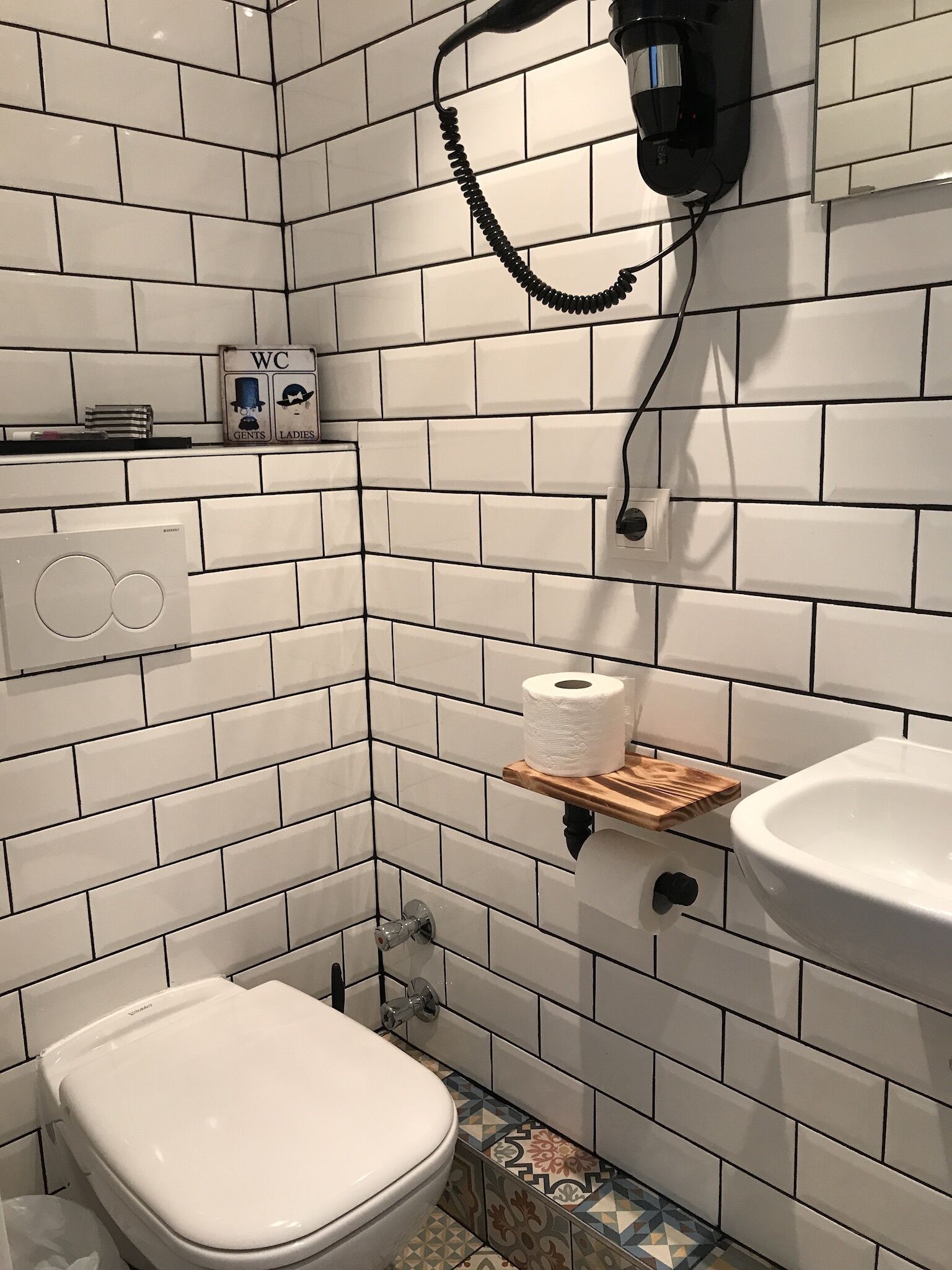 Une salle de bain avec des carreaux de céramique blancs rectangulaires, des toilettes, un lavabo et un sèche-mains fixé au mur.