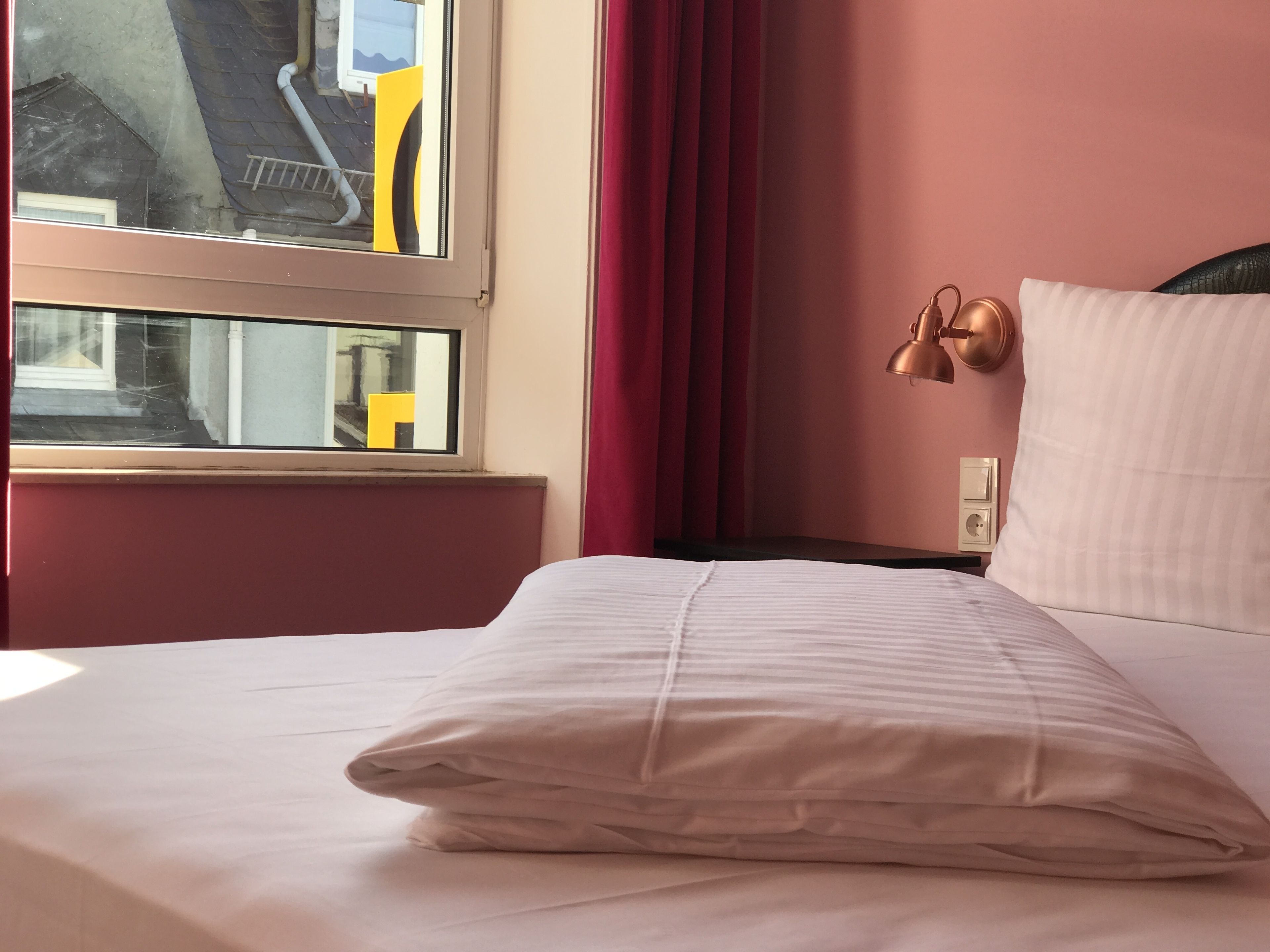 A neatly made bed with a white pillow and a striped blanket, a window with a view of a yellow ladder outside, and a wall-mounted lamp.